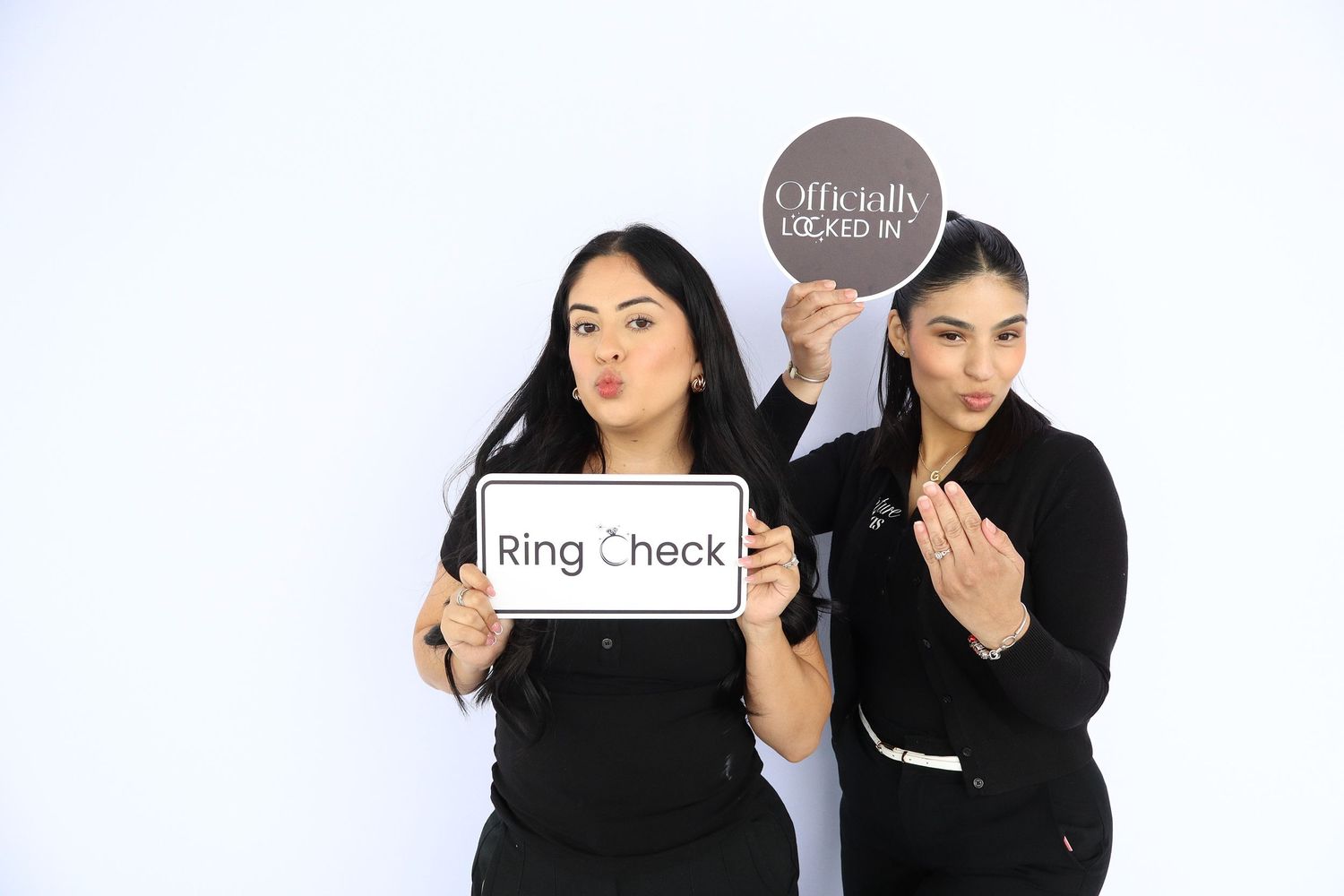 Two women in black outfits pose with Ring Check and Officially Booked In signs at a photo booth.