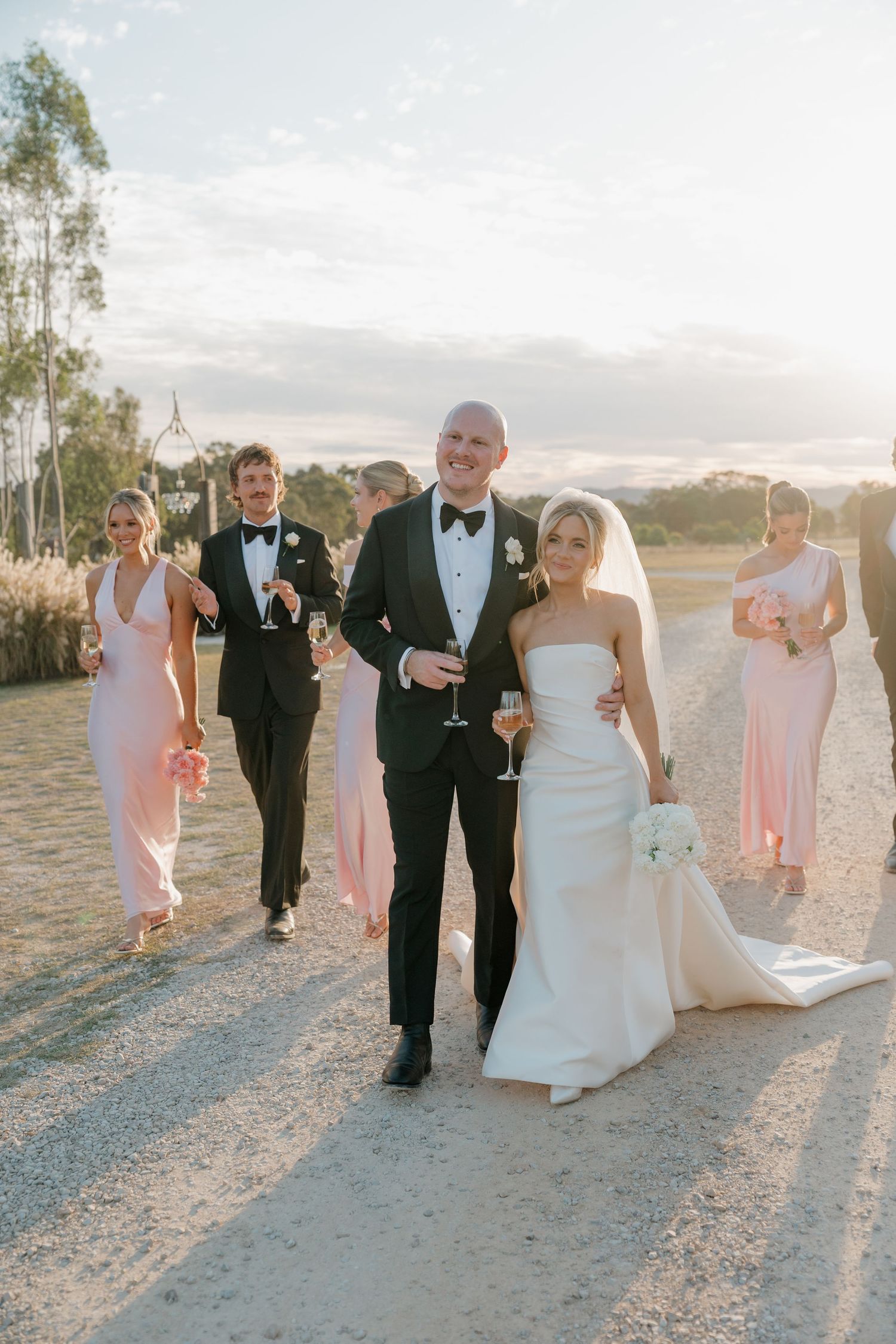 Wedding party walks outdoors at golden hour, bride in white gown with veil, groom in tuxedo, bridesmaids in blush pink dresses.