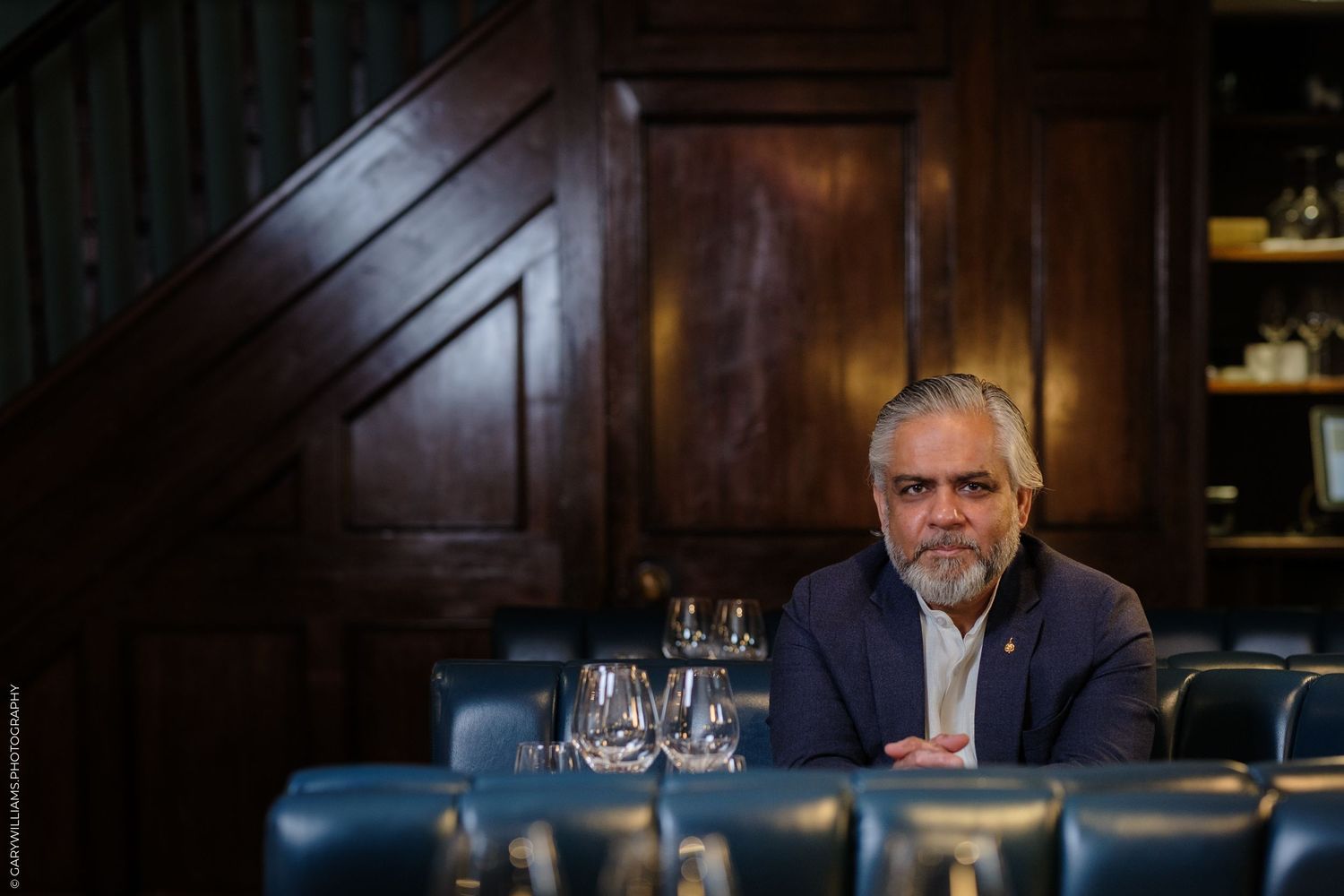 Distinguished man with beard sits at elegant restaurant table with wine glasses, dark wood paneling backdrop.