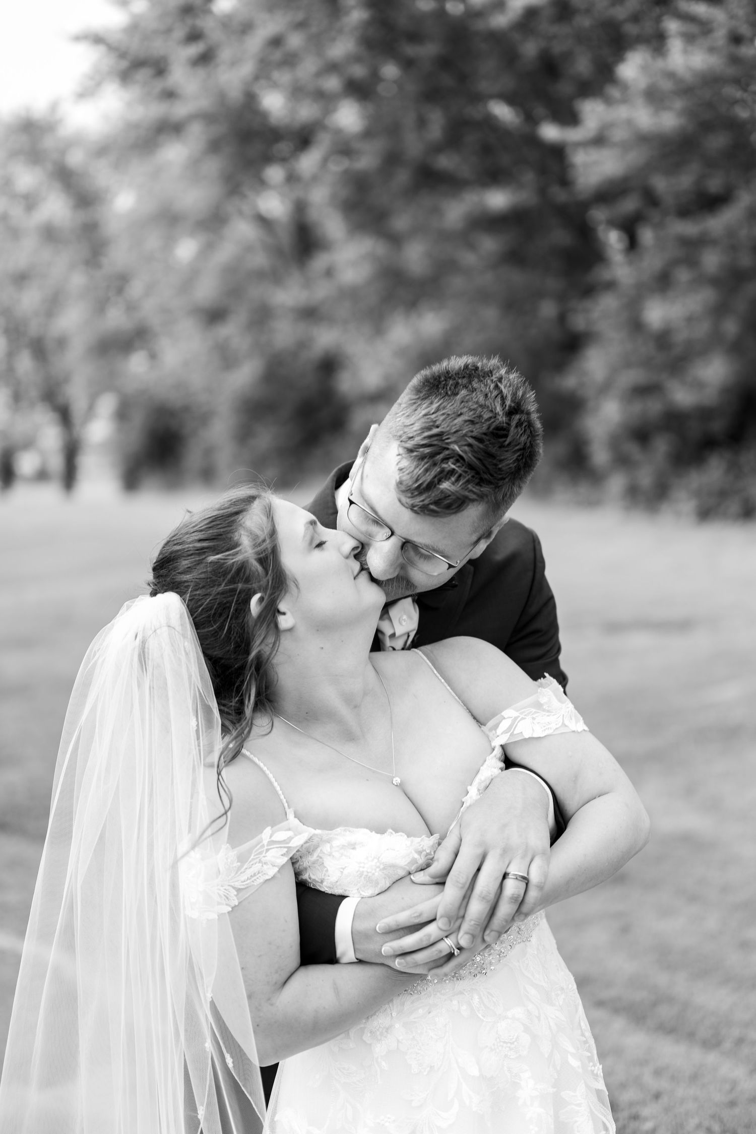 Black and white photo of a bride and groom sharing a romantic kiss outdoors, with the bride wearing a veil and lace gown.