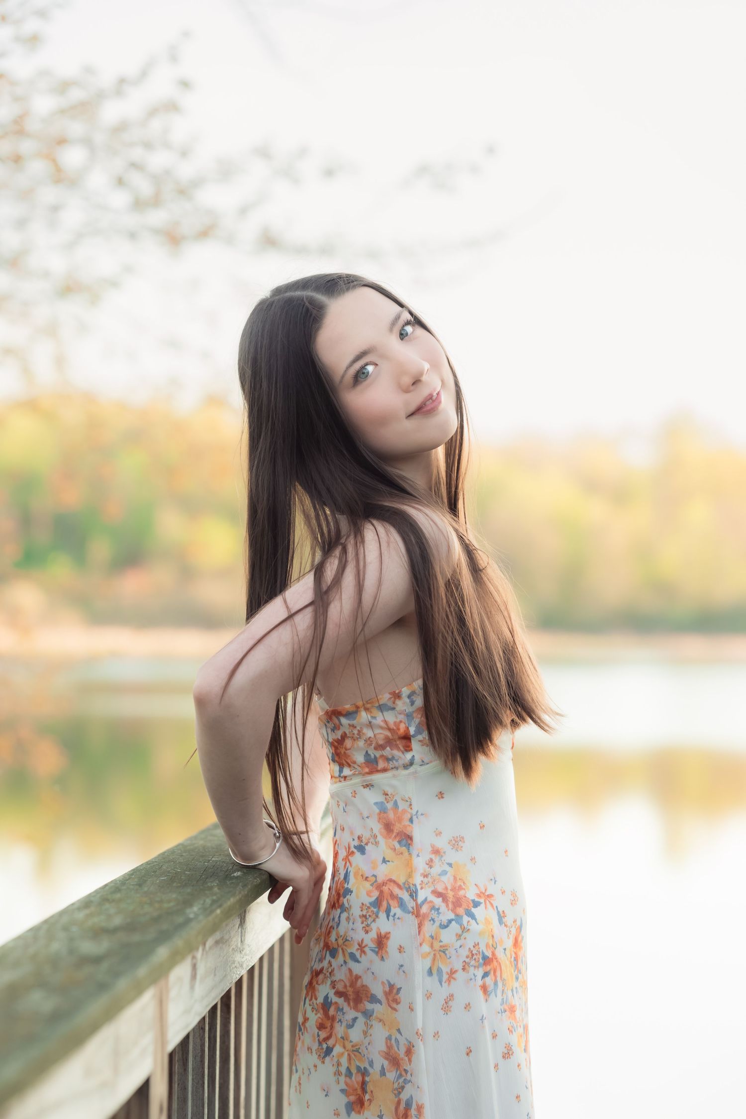 Young woman in floral sundress poses gracefully on wooden bridge overlooking serene lake during golden autumn afternoon.