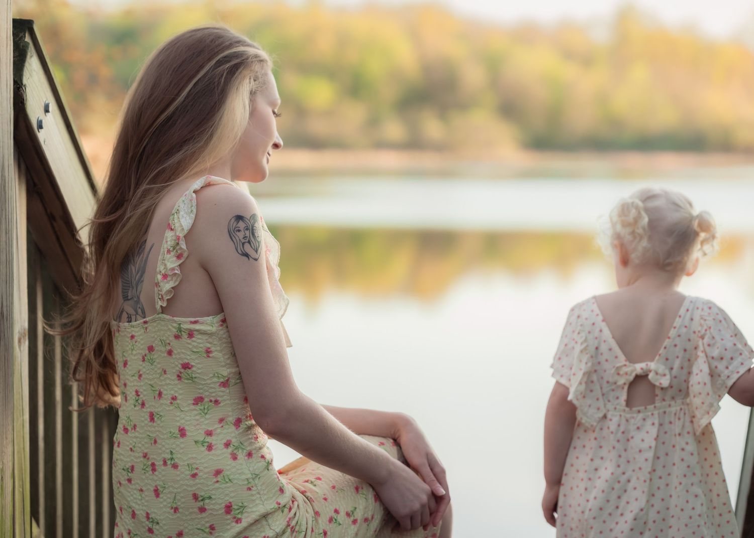 Two girls in floral dresses gaze over a serene lake surrounded by autumn trees in soft natural light.