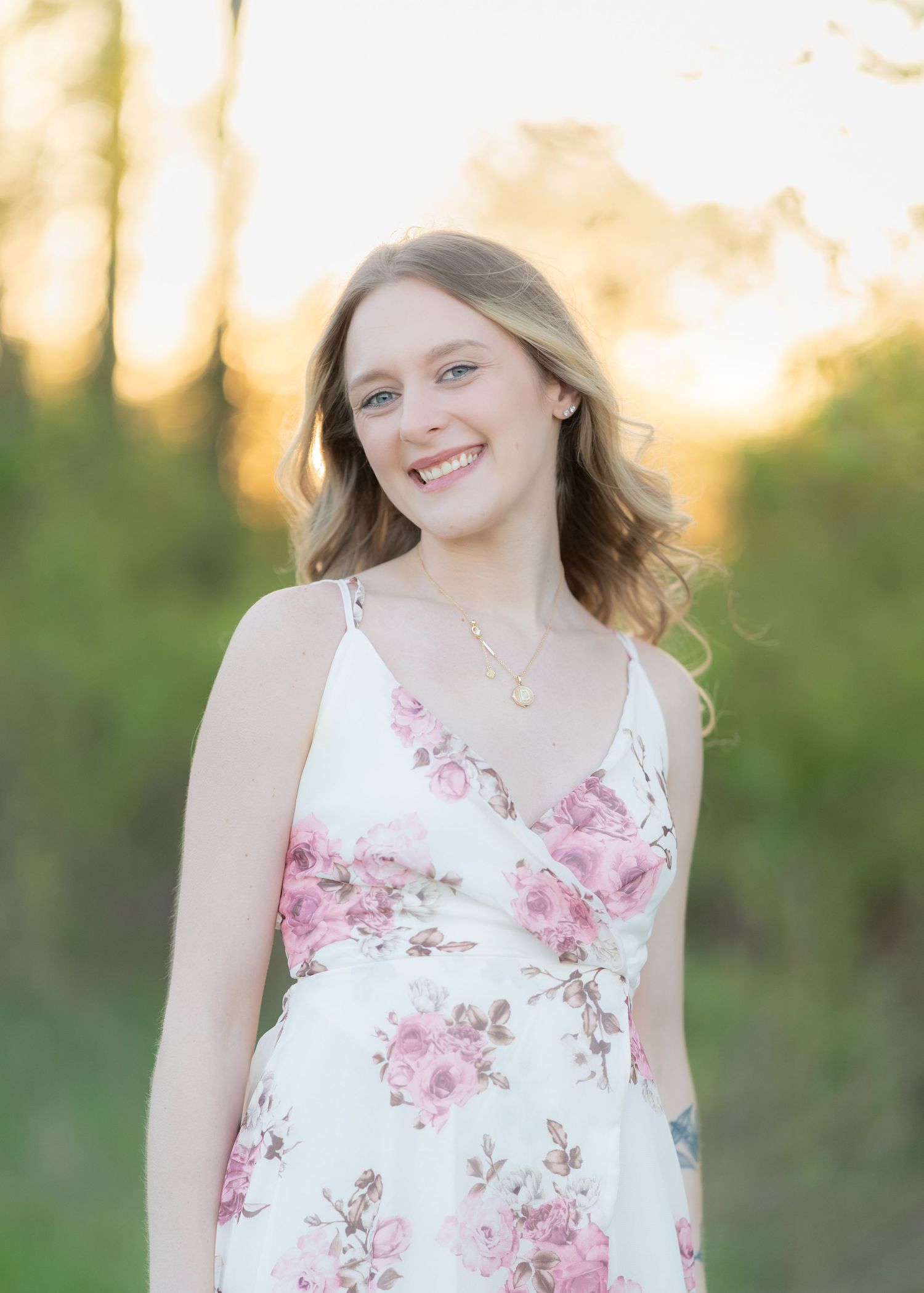 Smiling young woman in a floral white dress at golden hour outdoors.
