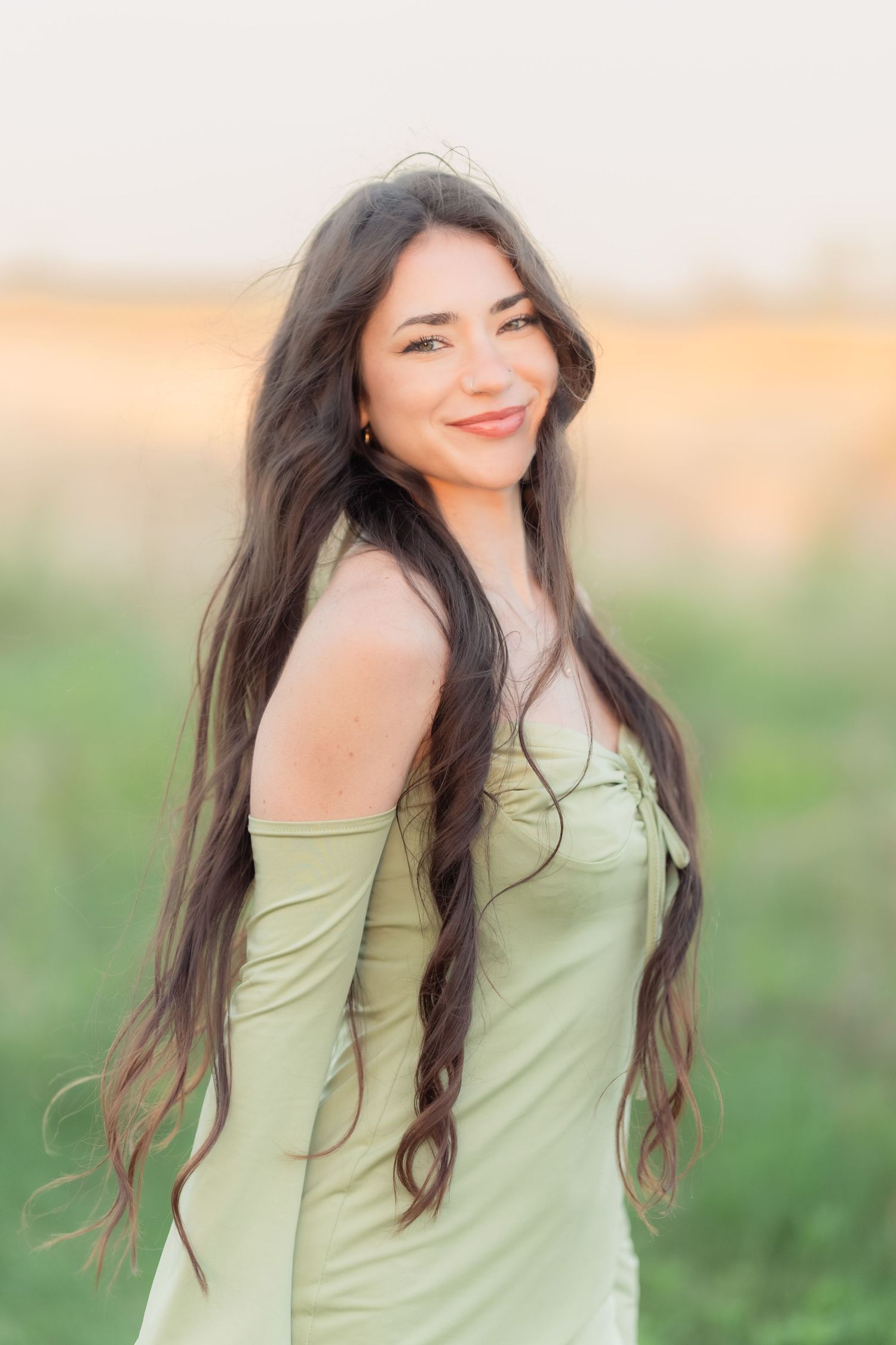 Young woman with long dark hair smiles outdoors in a sage green off-shoulder dress, golden hour light glowing behind her.