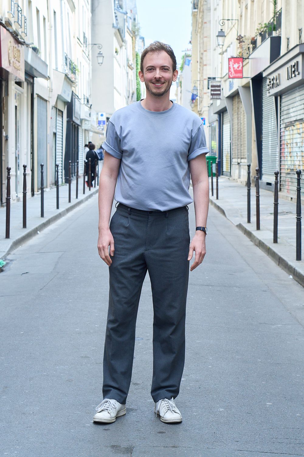 Man in blue t-shirt and gray trousers standing on a narrow European street, smiling at the camera.