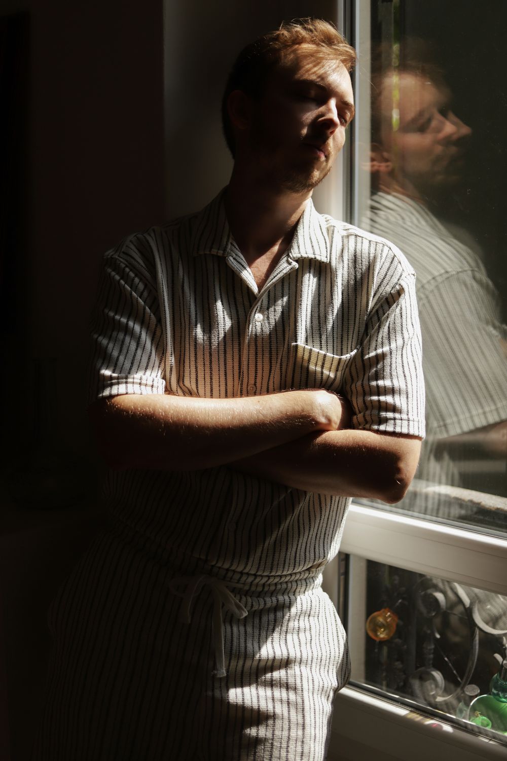 Man in striped shirt stands arms crossed by sunlit window with reflection visible in glass.