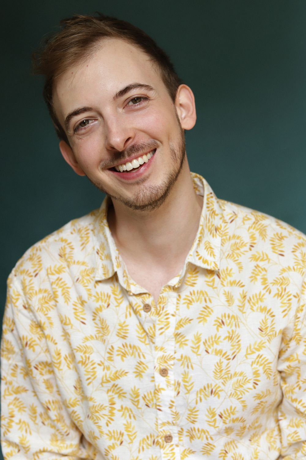 Smiling young man with light stubble wearing a floral yellow patterned shirt against a teal background.