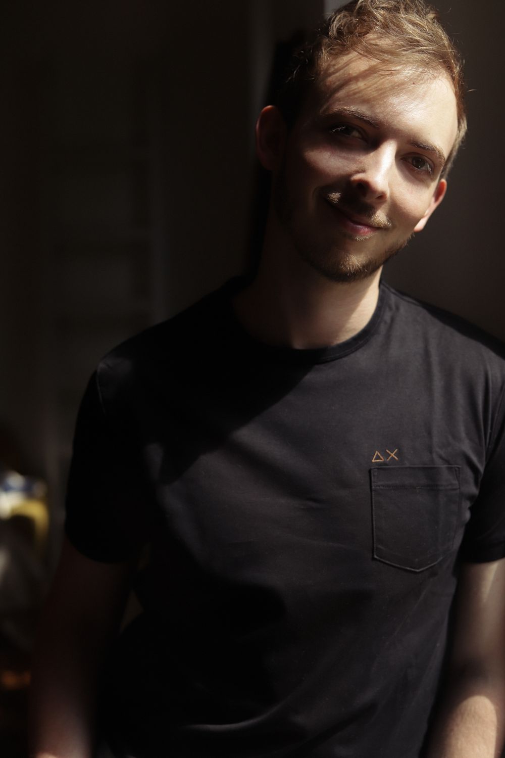Young man with light stubble smiling softly, wearing a black pocket t-shirt in dramatic moody lighting.