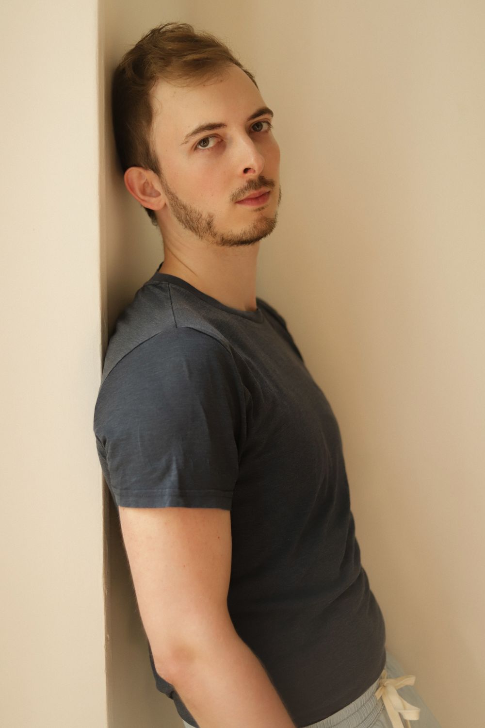 Young man with stubble leaning against a wall in a dark blue t-shirt, looking contemplatively upward.