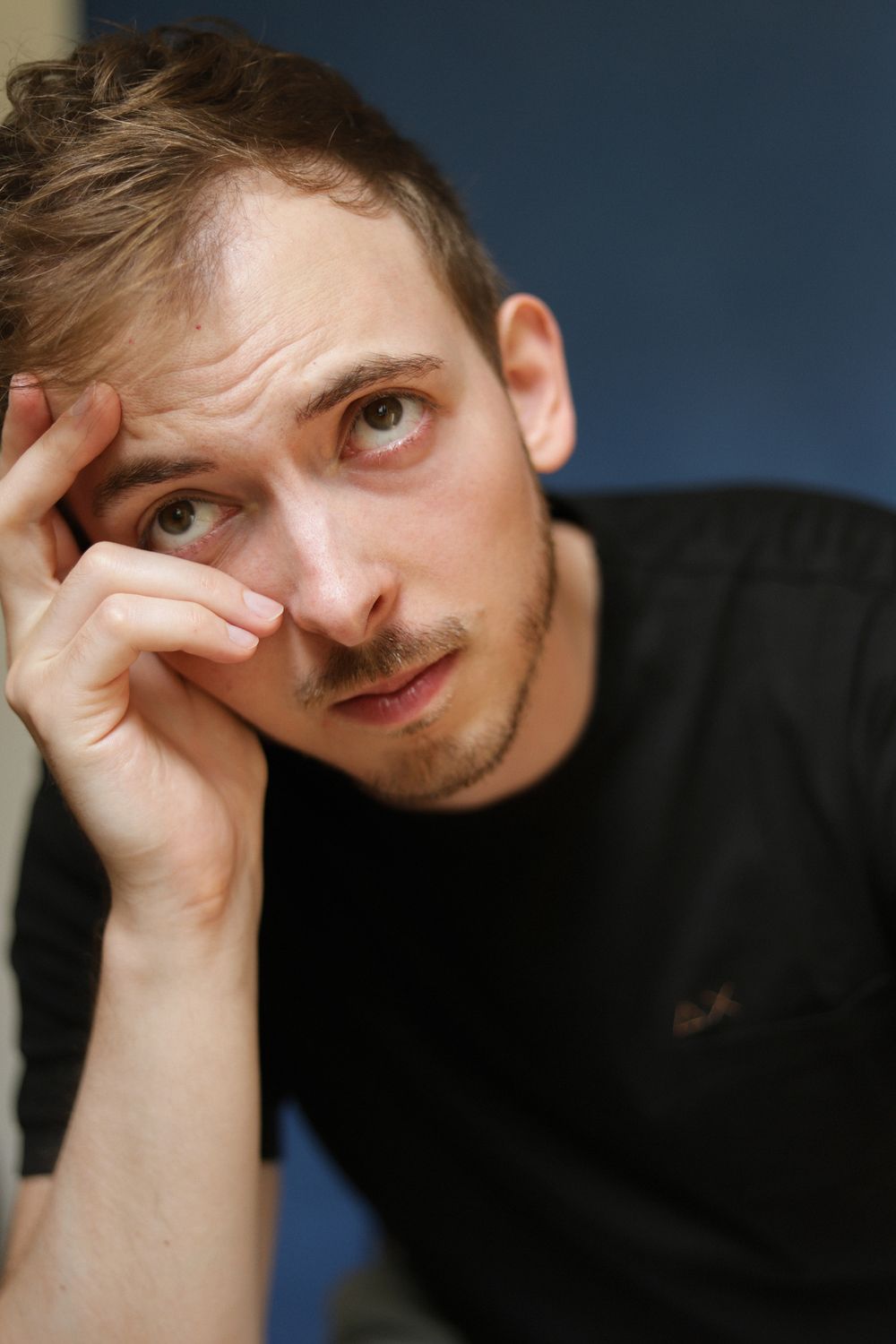 Young man in black t-shirt resting head on hand with thoughtful expression against blue background.