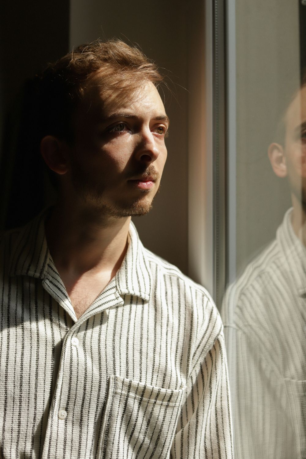 Young man in striped shirt gazing thoughtfully out a window with his reflection visible in the glass.