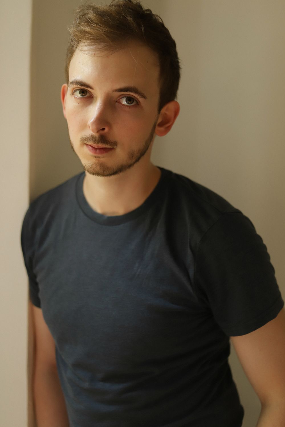 Young man with short brown hair and beard wearing a dark blue t-shirt, posing against a light wall.