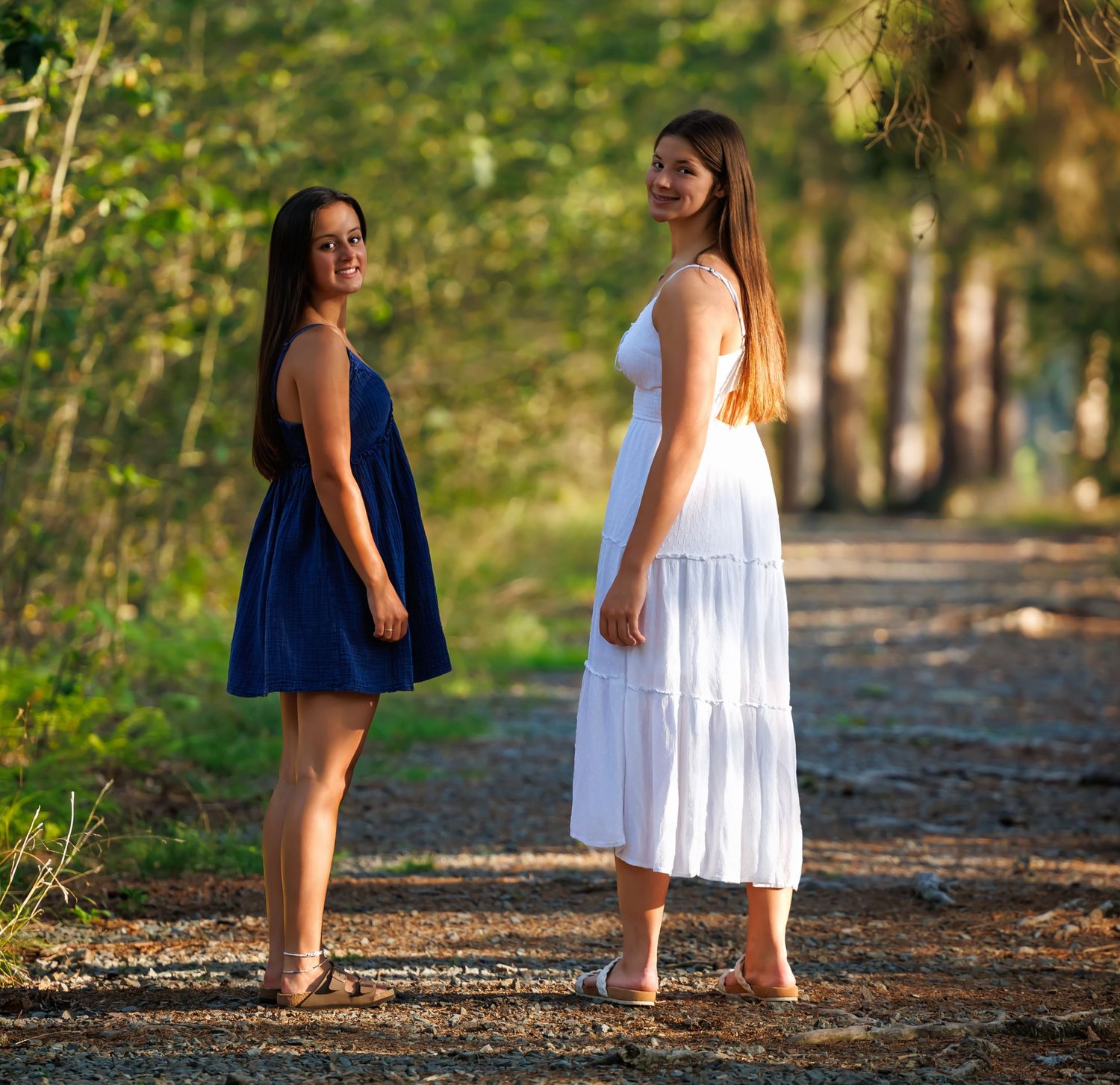 Two young women in blue and white dresses stand on a sunlit wooded path smiling.