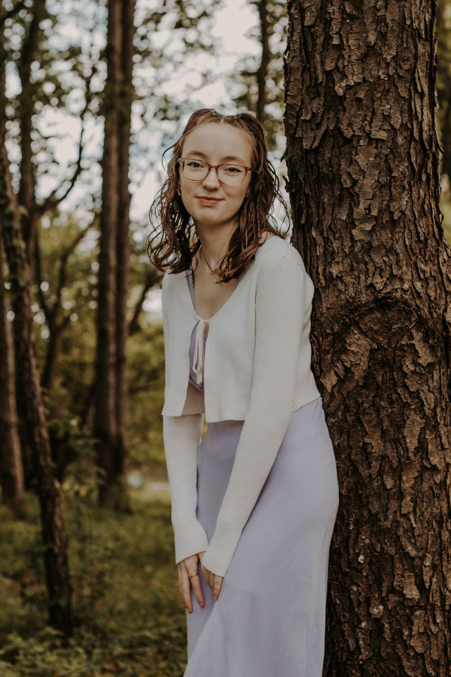 Young woman in white cardigan and lavender skirt leaning against a tree in a lush forest setting.