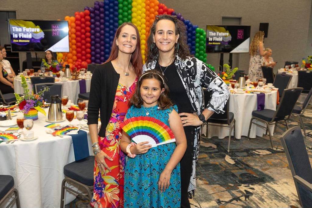 Two women and a young girl pose at a colorful Pride event with rainbow balloon arch and banquet tables.