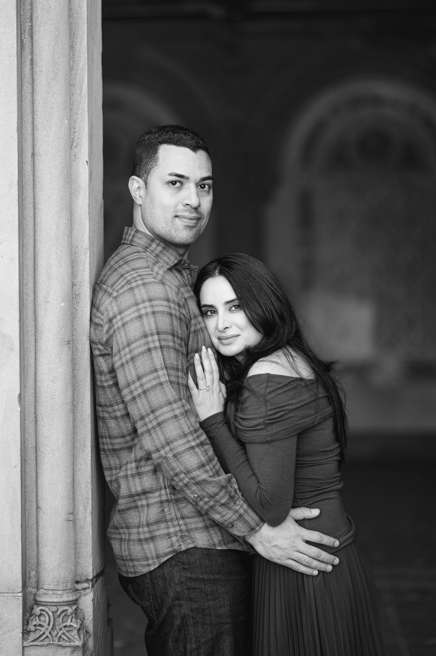 Black and white photo of a couple embracing by an ornate column, woman in off-shoulder dress, man in plaid shirt.