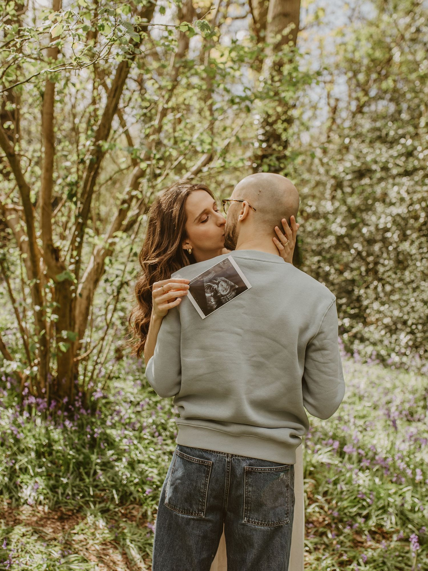 Couple kissing in a woodland garden holding an ultrasound scan photo for a pregnancy announcement.
