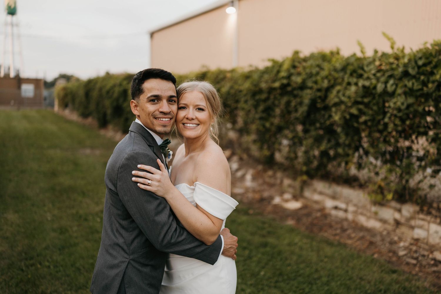 Smiling bride and groom embracing outdoors near a green hedge at their wedding.