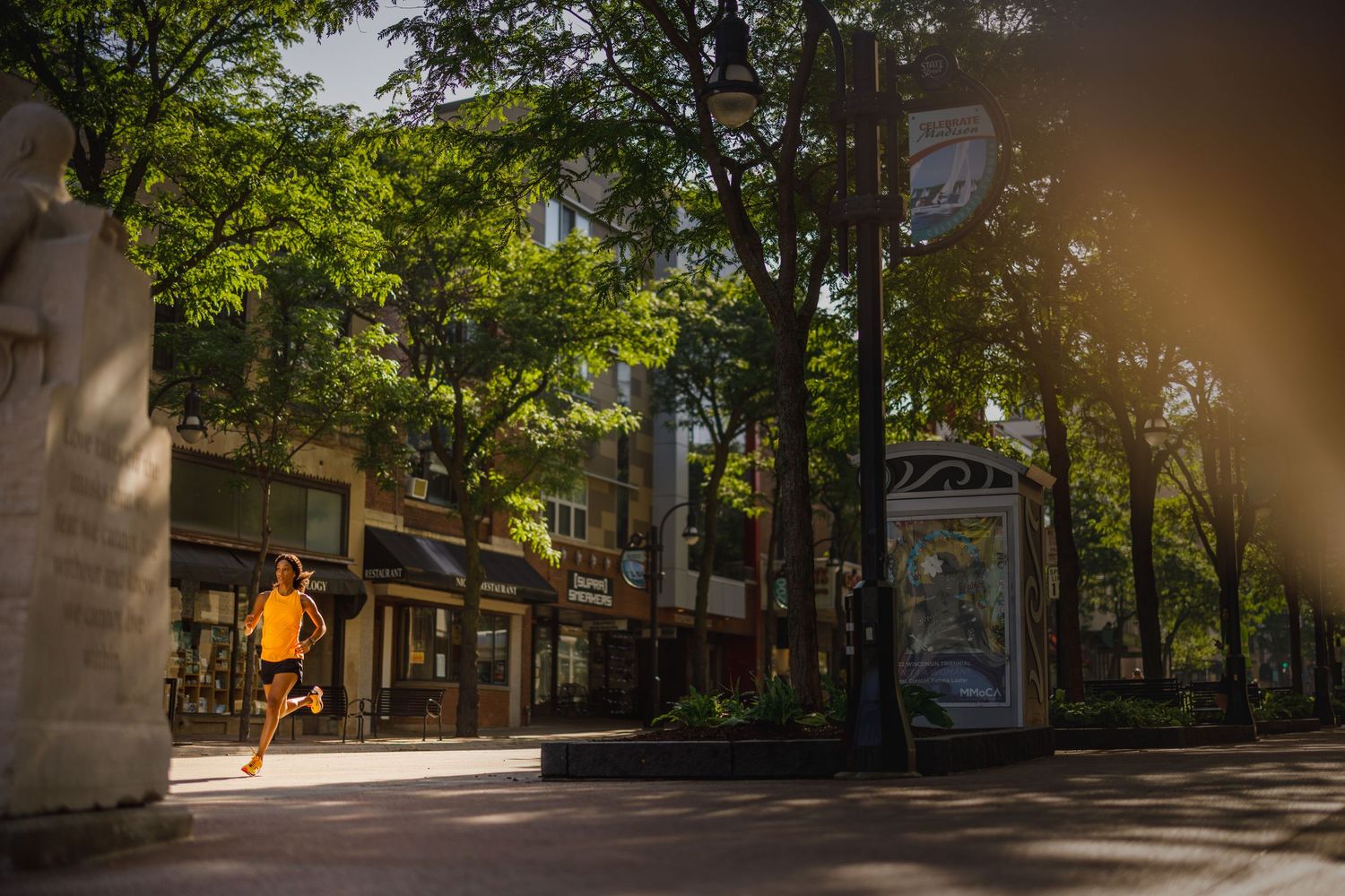 A jogger in orange runs through a tree-lined urban pedestrian plaza bathed in warm golden sunlight.