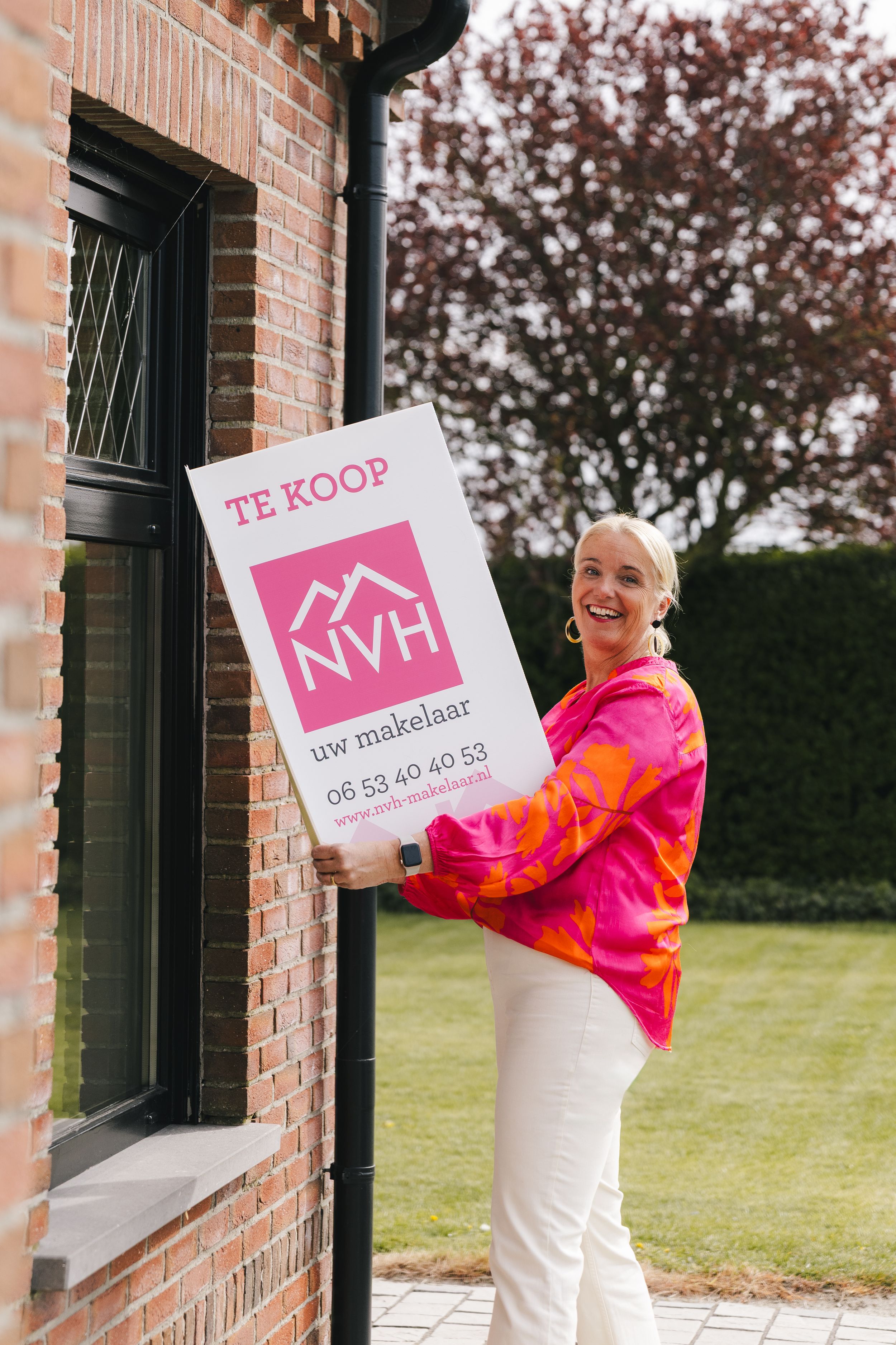 Smiling blonde woman holding a pink NVH real estate for sale sign outside a brick house.