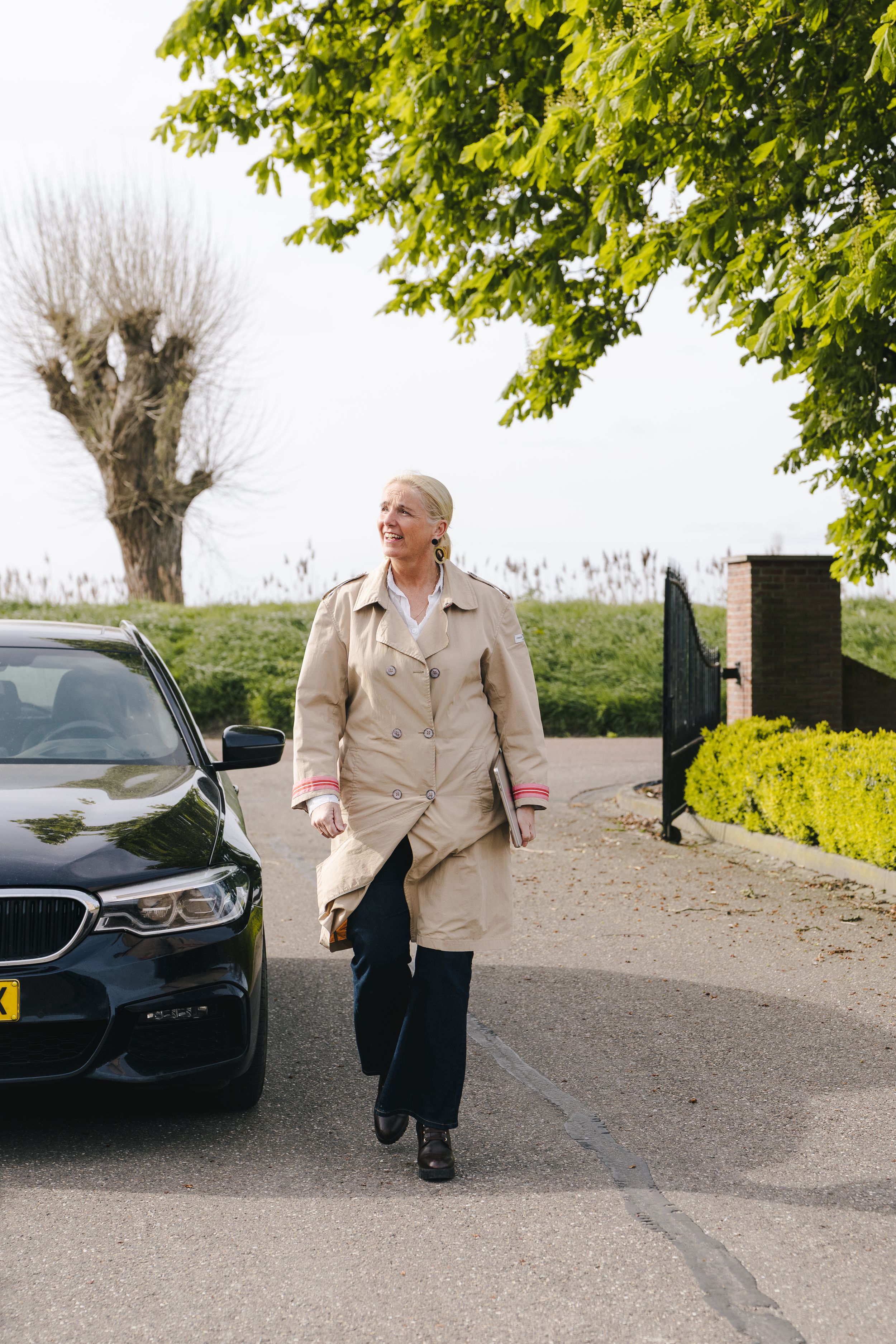 Woman in beige trench coat walking beside a black BMW on a paved driveway with green trees in background.