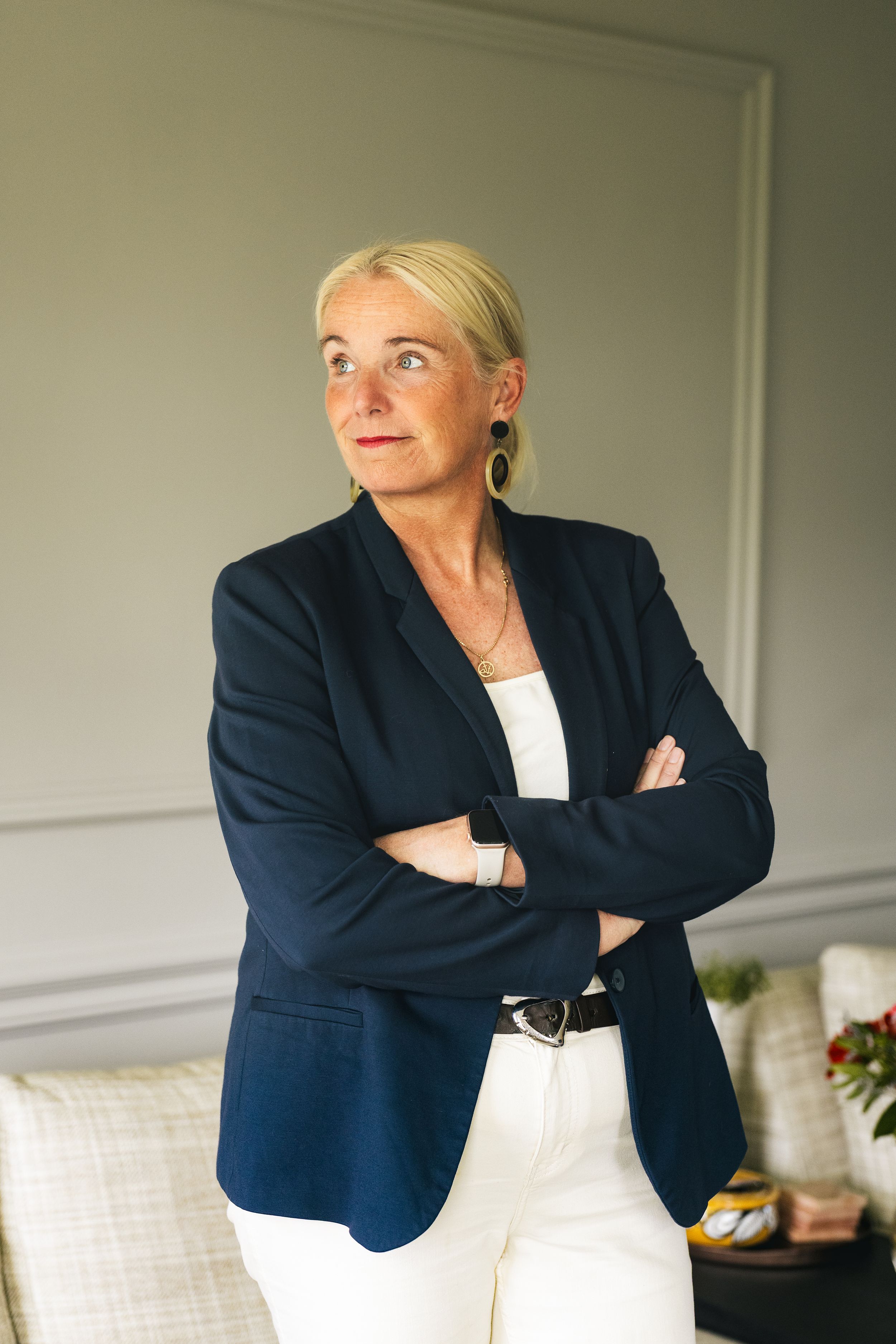 Professional blonde woman in navy blazer and white pants stands with arms crossed in elegant interior.