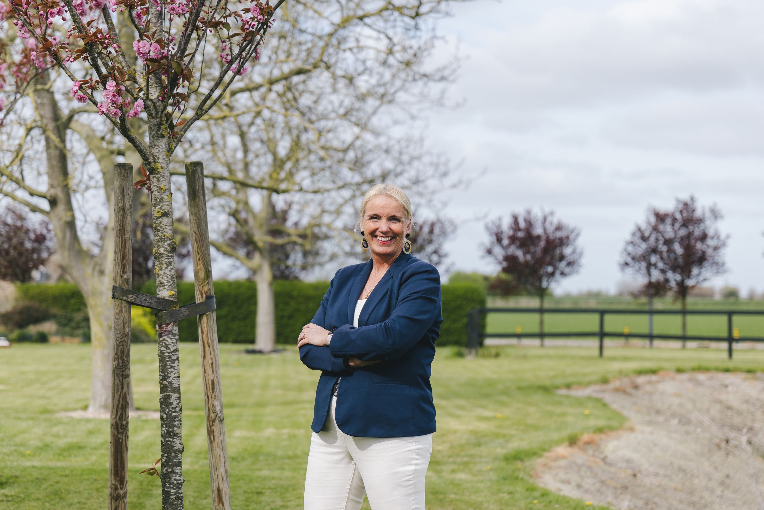 Woman in navy blazer and white pants smiling outdoors near blooming trees and wooden fence.