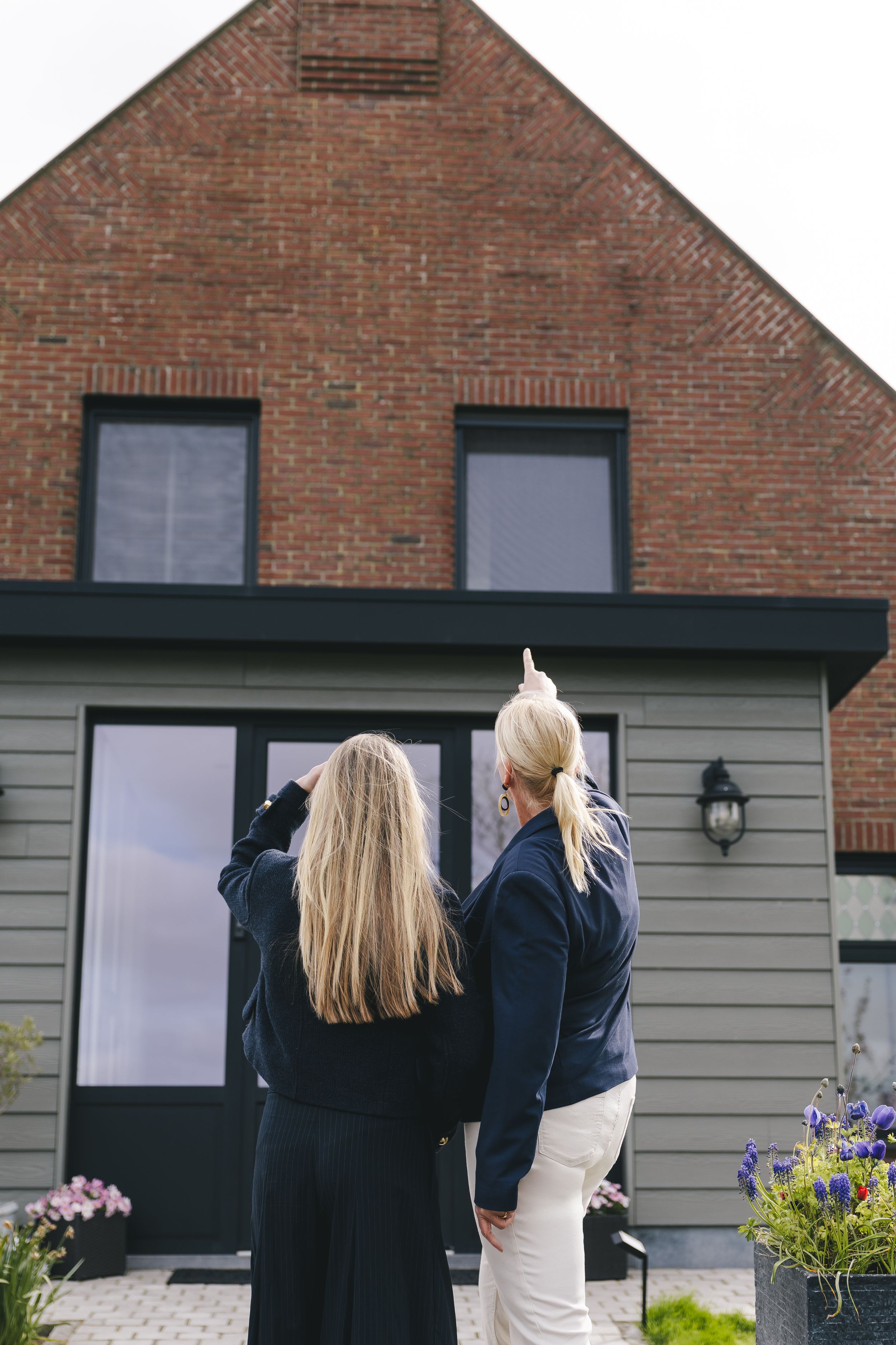 Two women with blonde hair stand outside a modern brick house, one pointing upward while viewing the property.