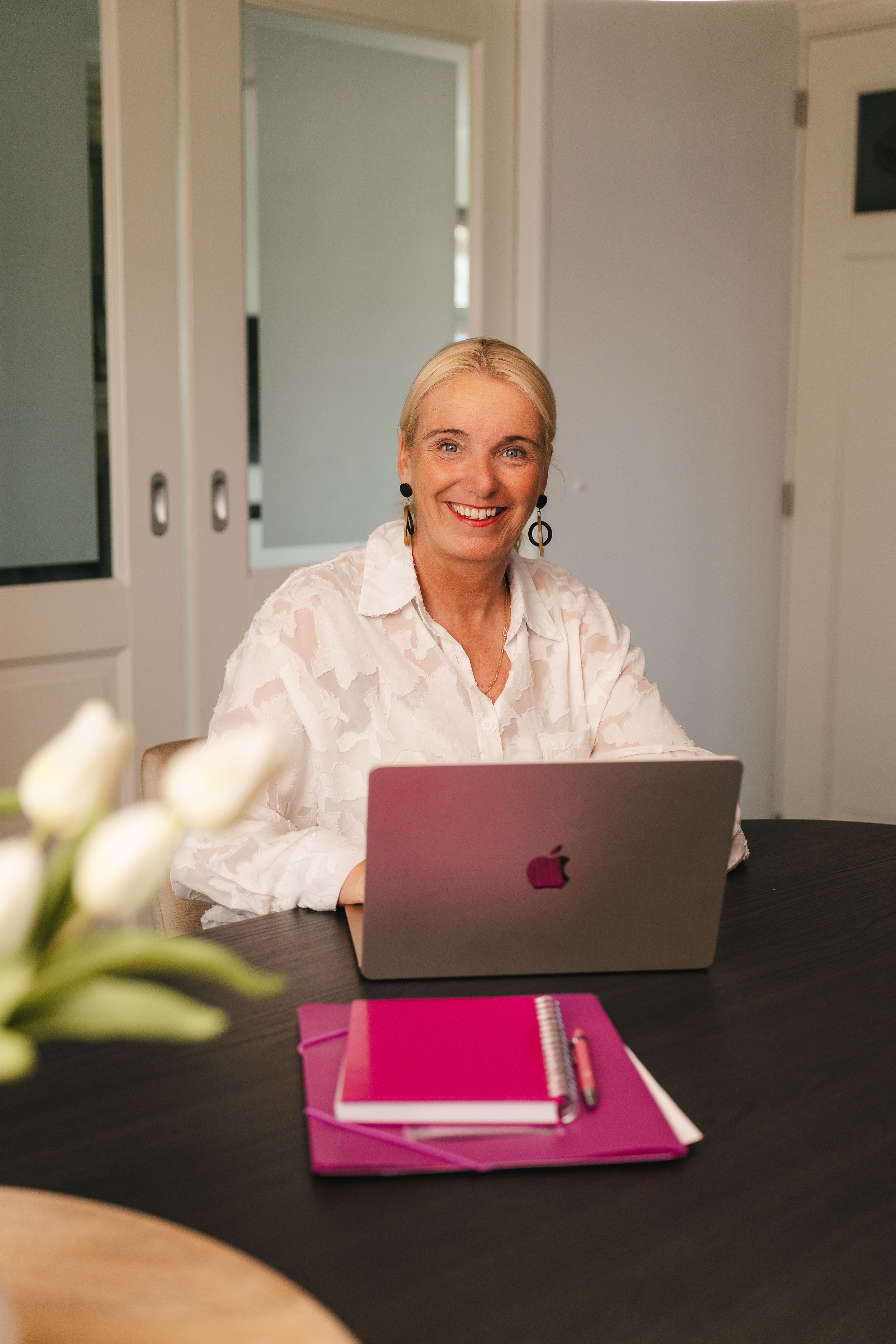 Smiling blonde woman sitting at a dark desk with a MacBook laptop, pink notebook, and white tulips in a bright room.