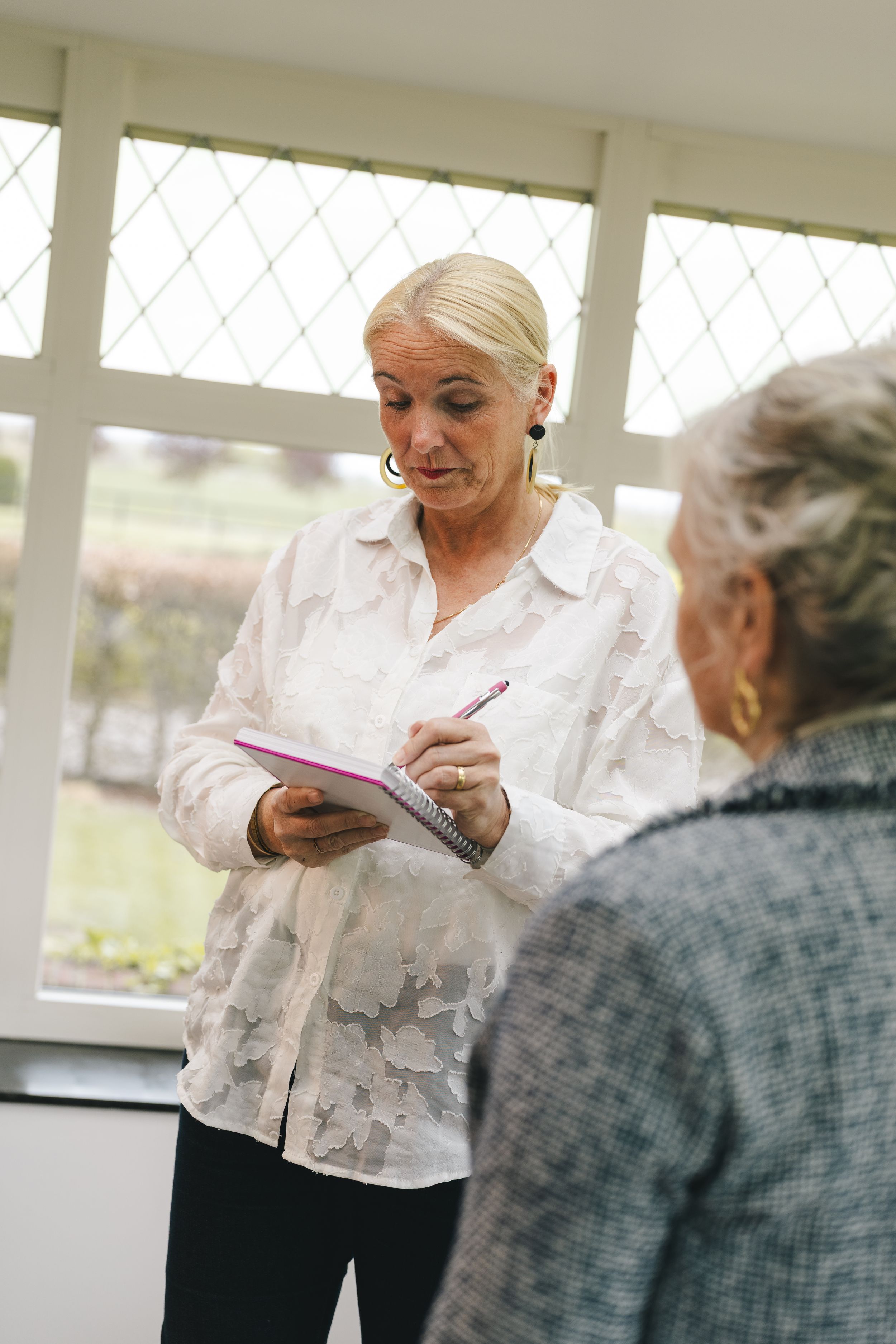 A healthcare worker with a clipboard consults an elderly woman in a bright, windowed room.