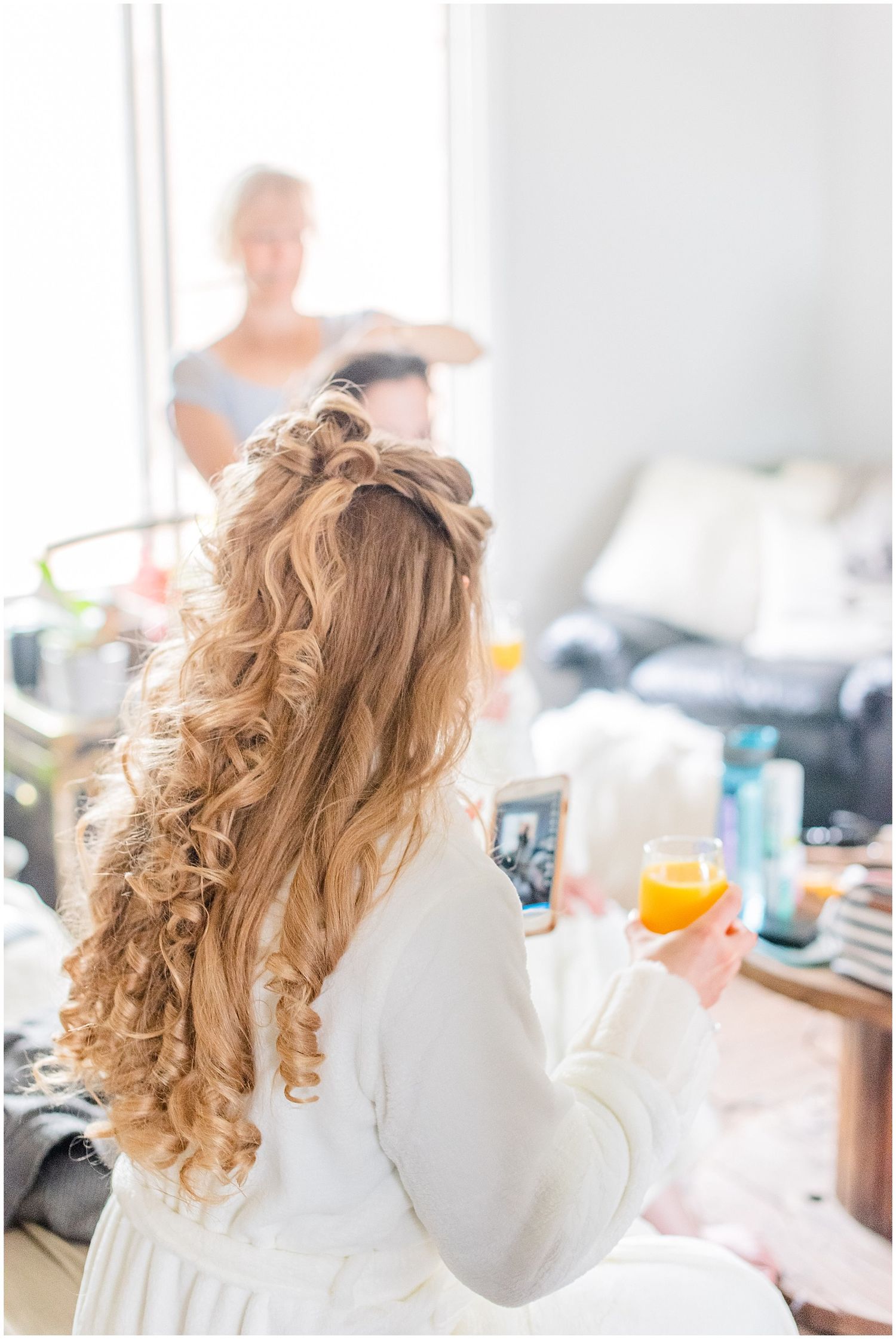 Bride in white robe with curly half-up hairstyle holding orange juice and phone while getting ready.