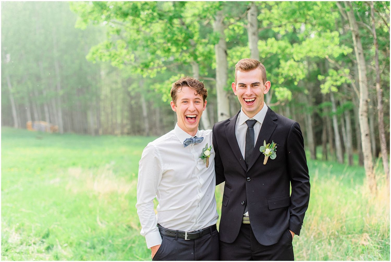 Two smiling groomsmen in black suits with boutonnieres laugh together in a green outdoor setting.