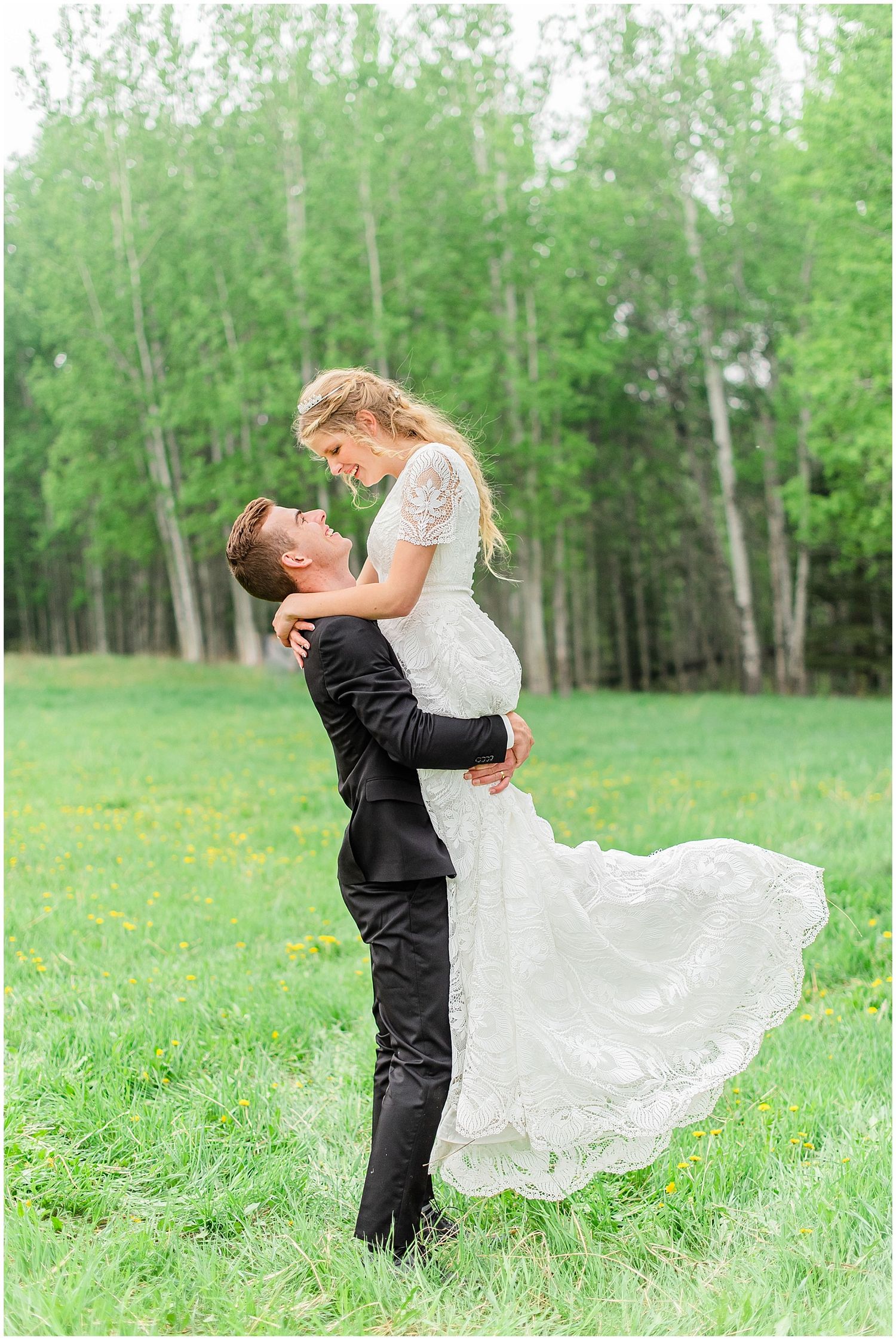 Groom lifts bride in white lace wedding dress in a lush green meadow with birch trees.