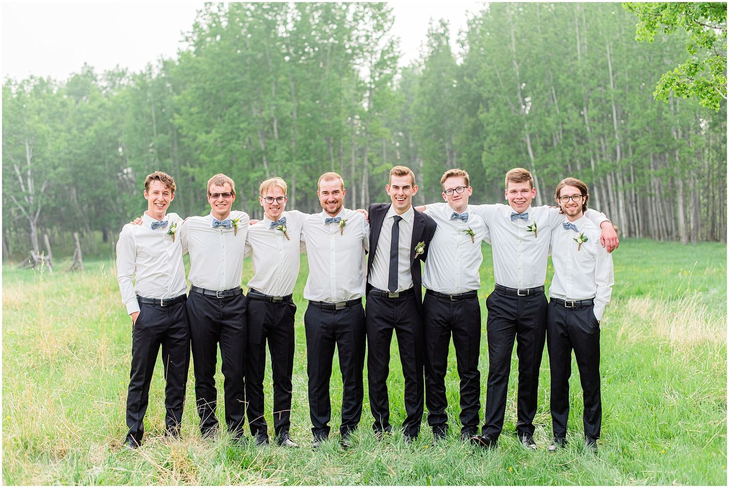 A groom and his seven groomsmen pose in white shirts and black pants in a lush green field.