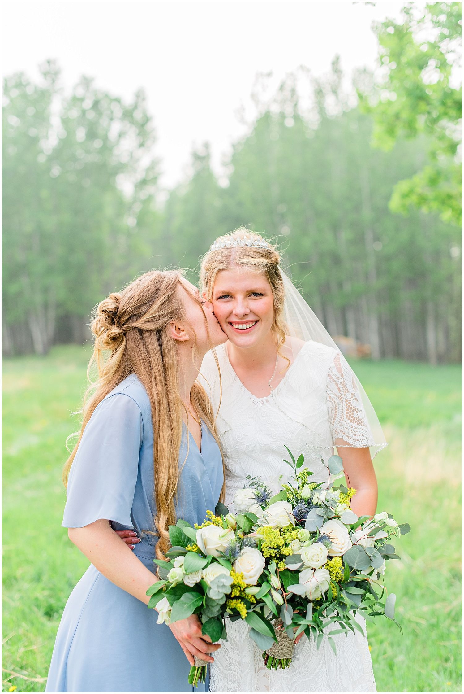 Bridesmaid in blue dress kisses smiling bride holding white floral bouquet.