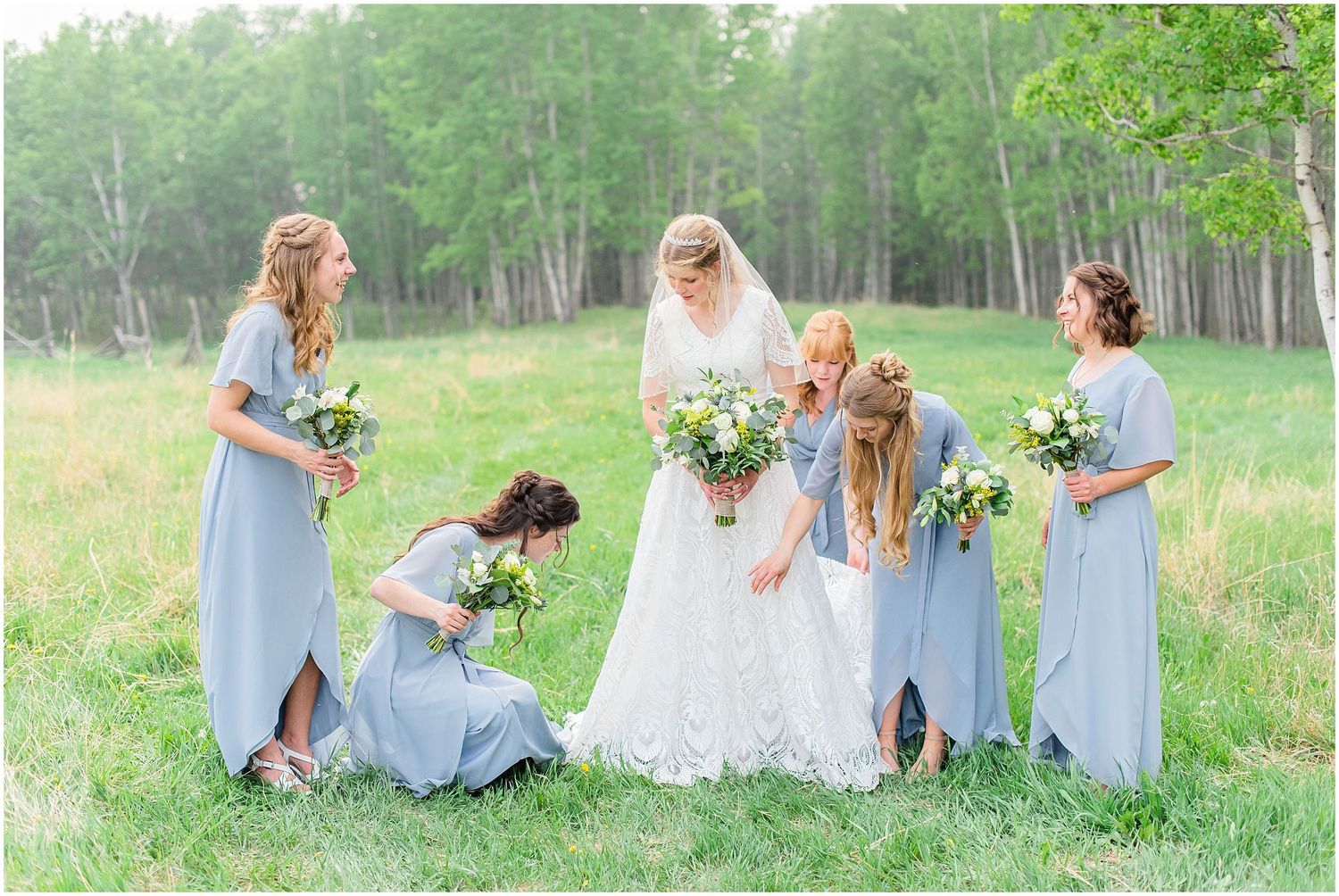 Bride surrounded by bridesmaids in dusty blue dresses in a green meadow.