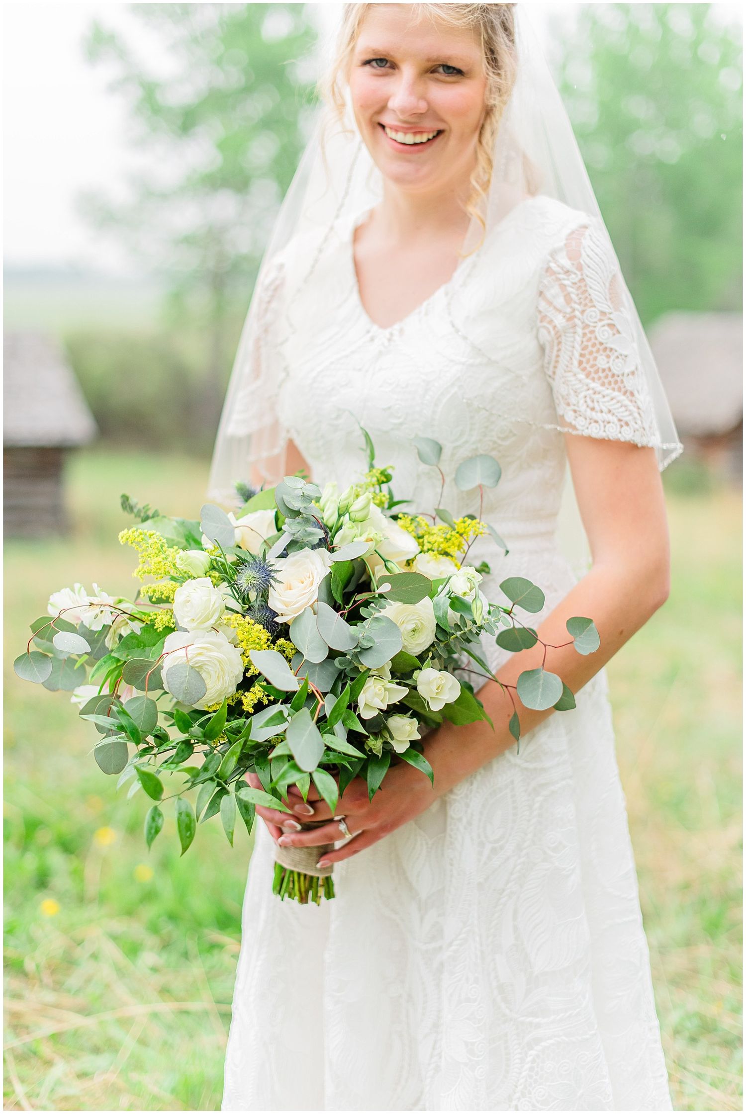 A smiling bride in a lace wedding dress holds a bouquet of white roses and greenery outdoors.