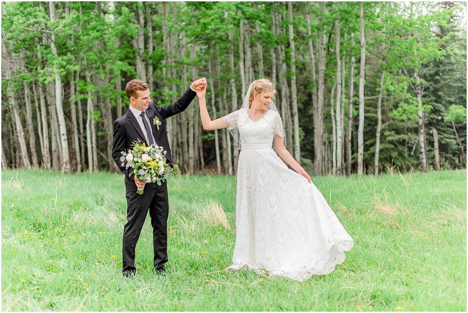 Groom twirls bride in white lace gown in a green meadow surrounded by tall trees on wedding day.