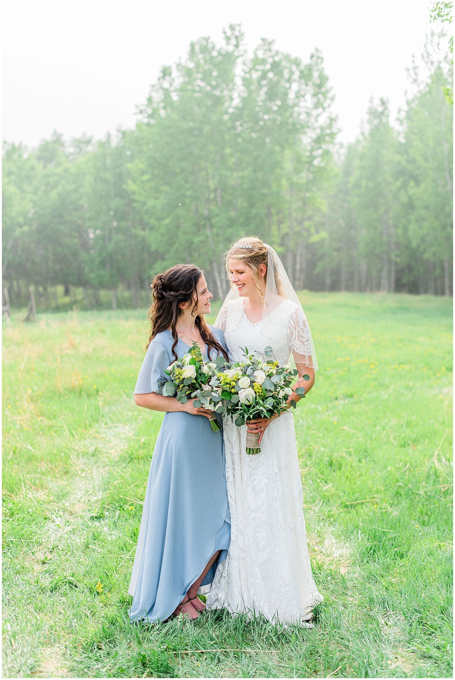 Bride in lace gown and bridesmaid in blue dress share a joyful moment in a green field with bouquet.