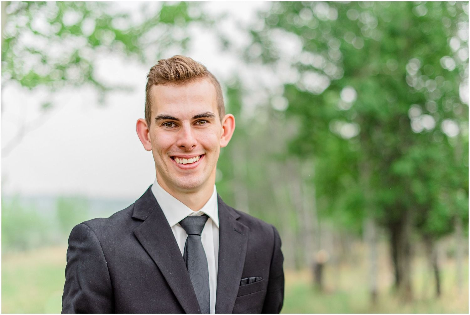 Handsome groom in dark suit and gray tie smiling outdoors with trees in soft focus background.