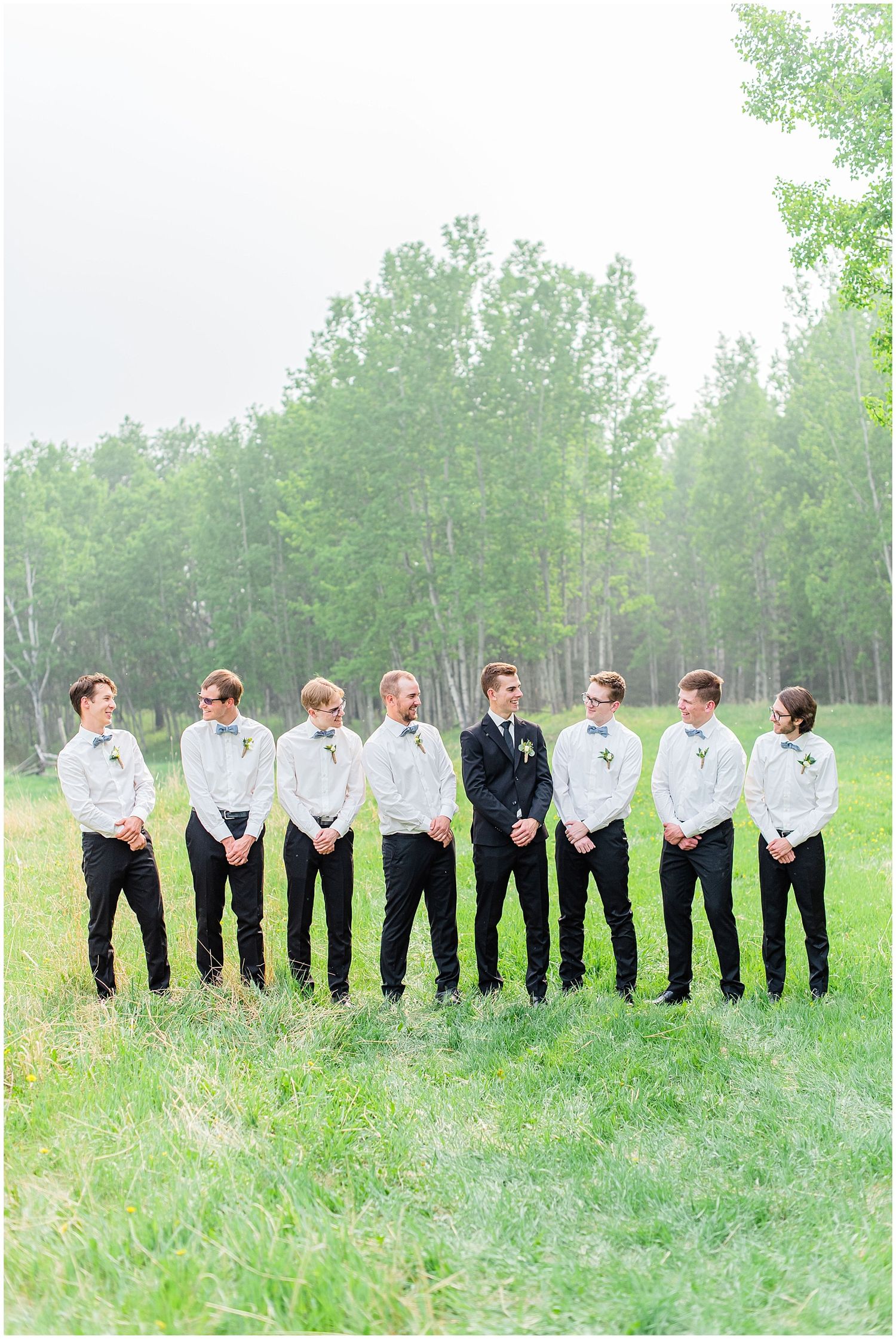 A groom in a black suit stands with seven groomsmen in white shirts on a green grass field.