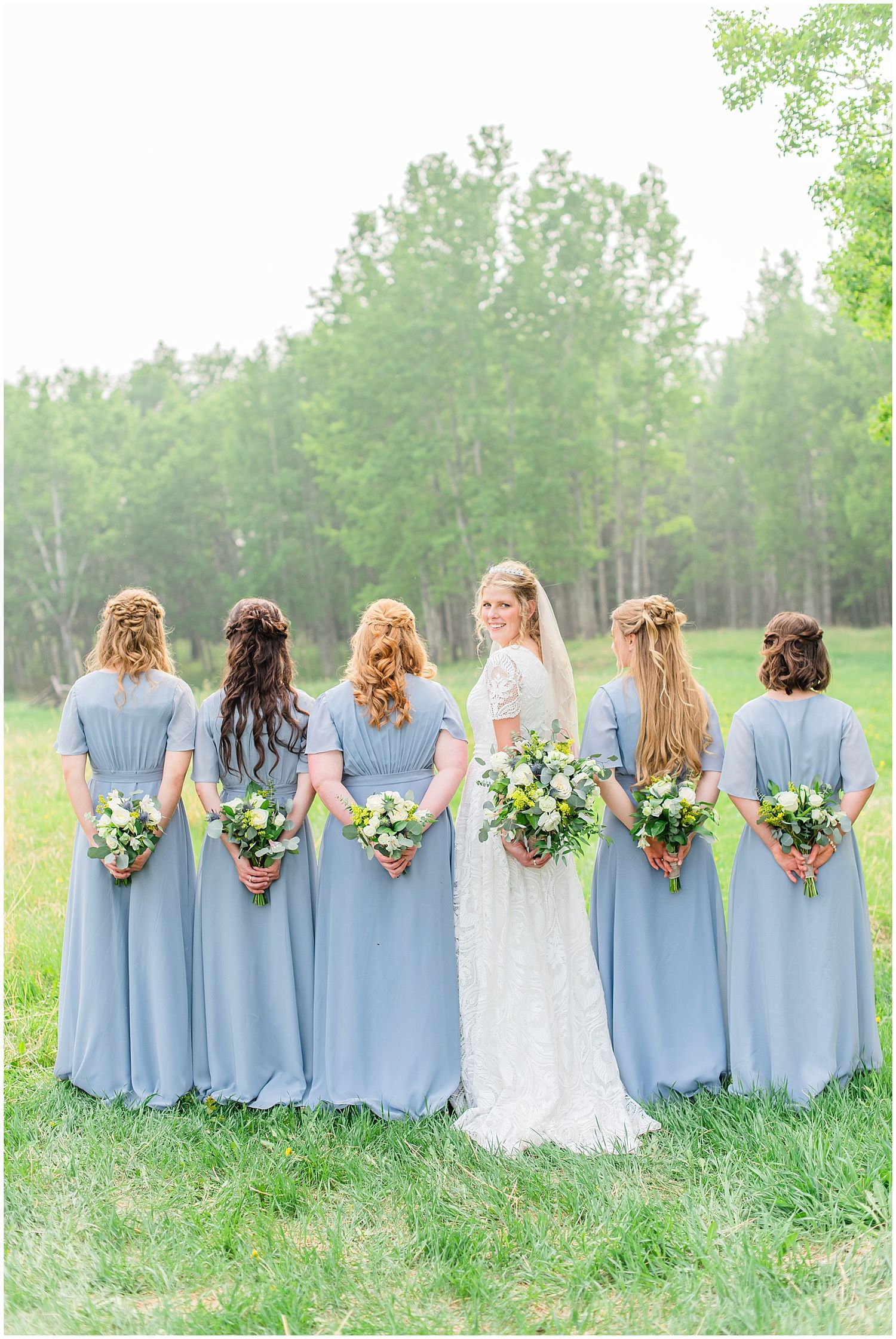 Bride in white lace gown stands with six bridesmaids in dusty blue dresses, all facing away holding bouquets.