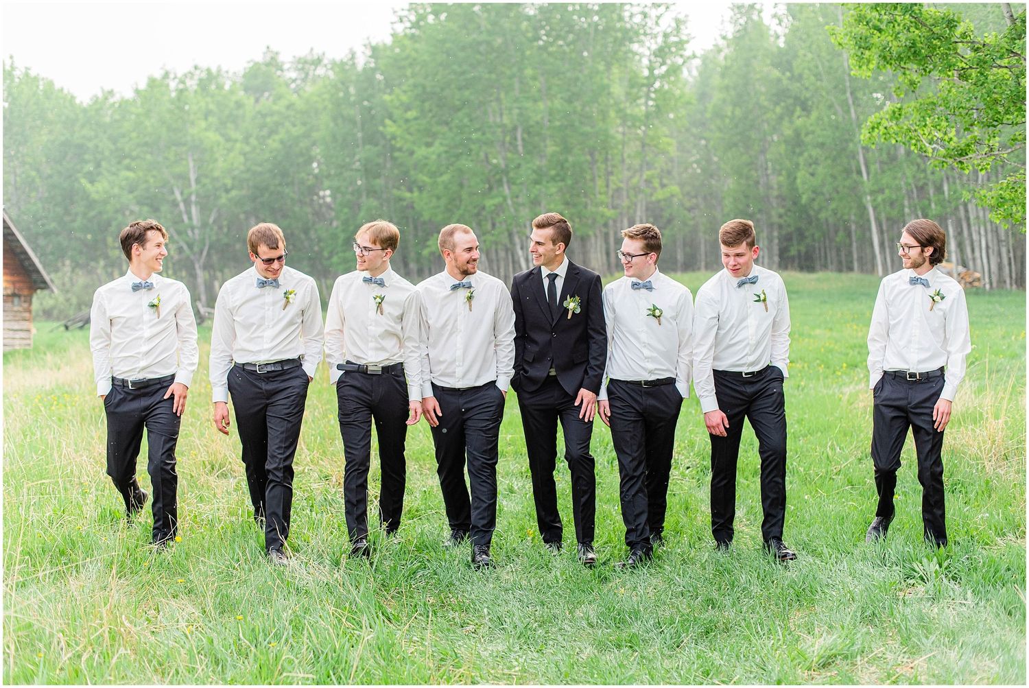 Groom in black suit stands confidently with seven groomsmen in white shirts on green grass field.