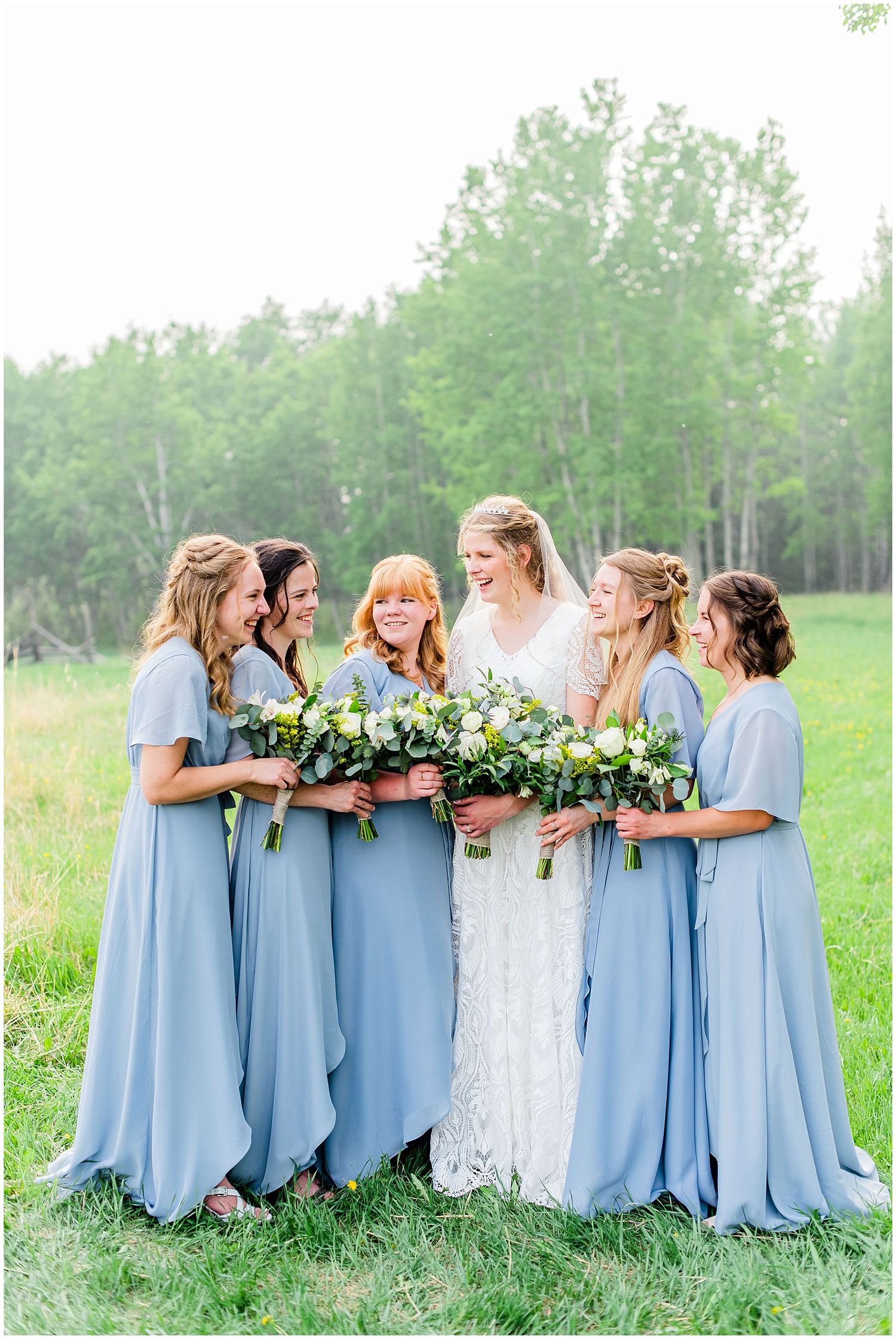 Bride surrounded by bridesmaids in dusty blue dresses holding white and green bouquets outdoors.