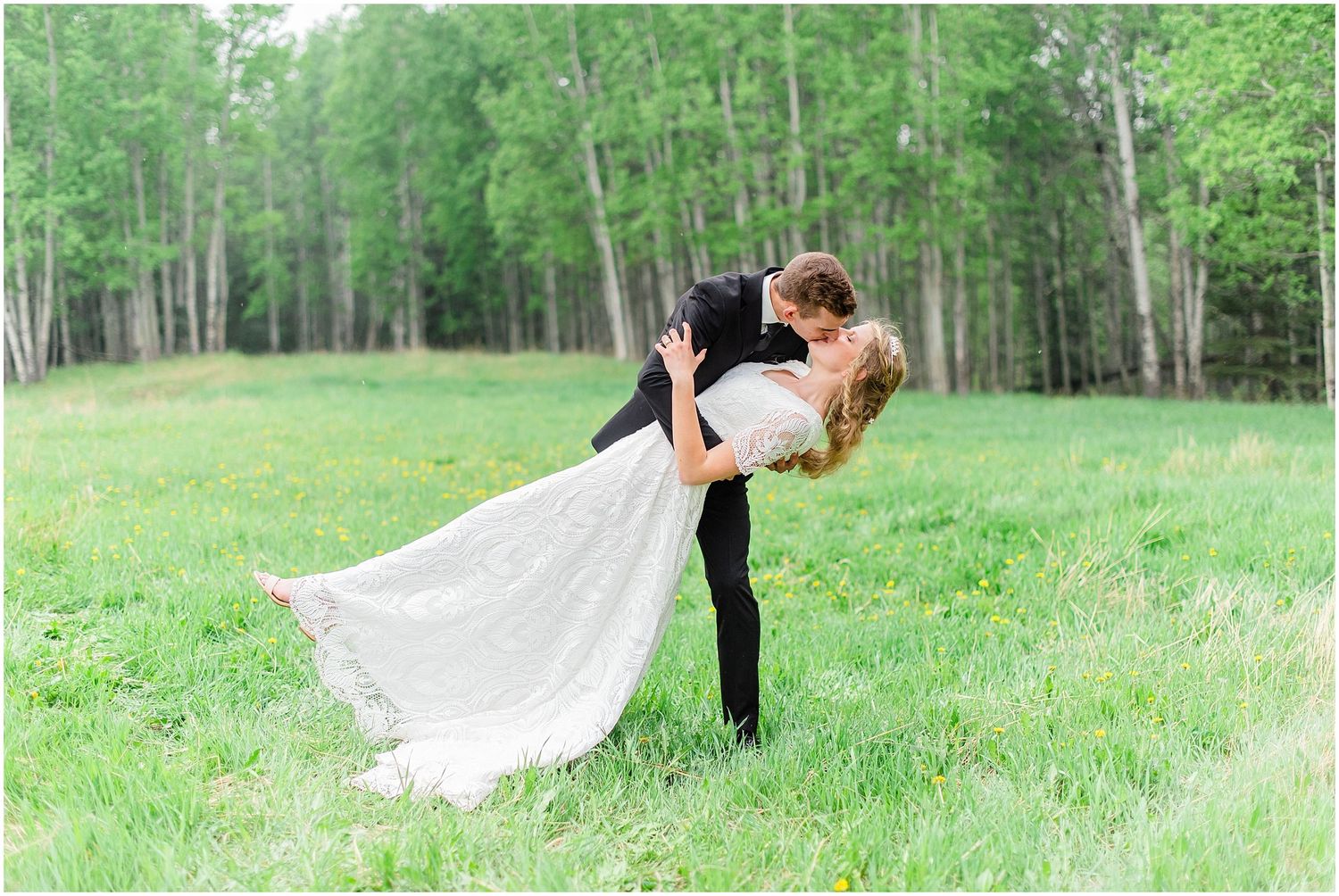 Groom dips bride in romantic pose in lush green meadow surrounded by trees.