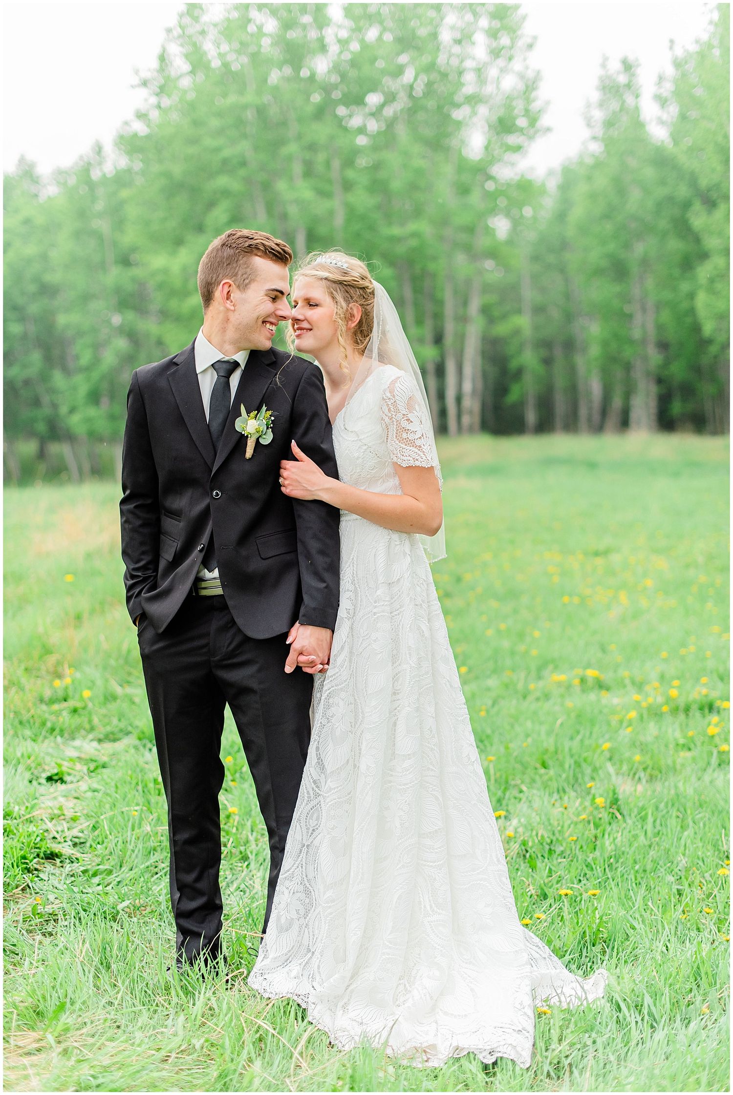 Bride and groom share a romantic moment in a lush green field, bride wearing lace gown with veil, groom in black suit.