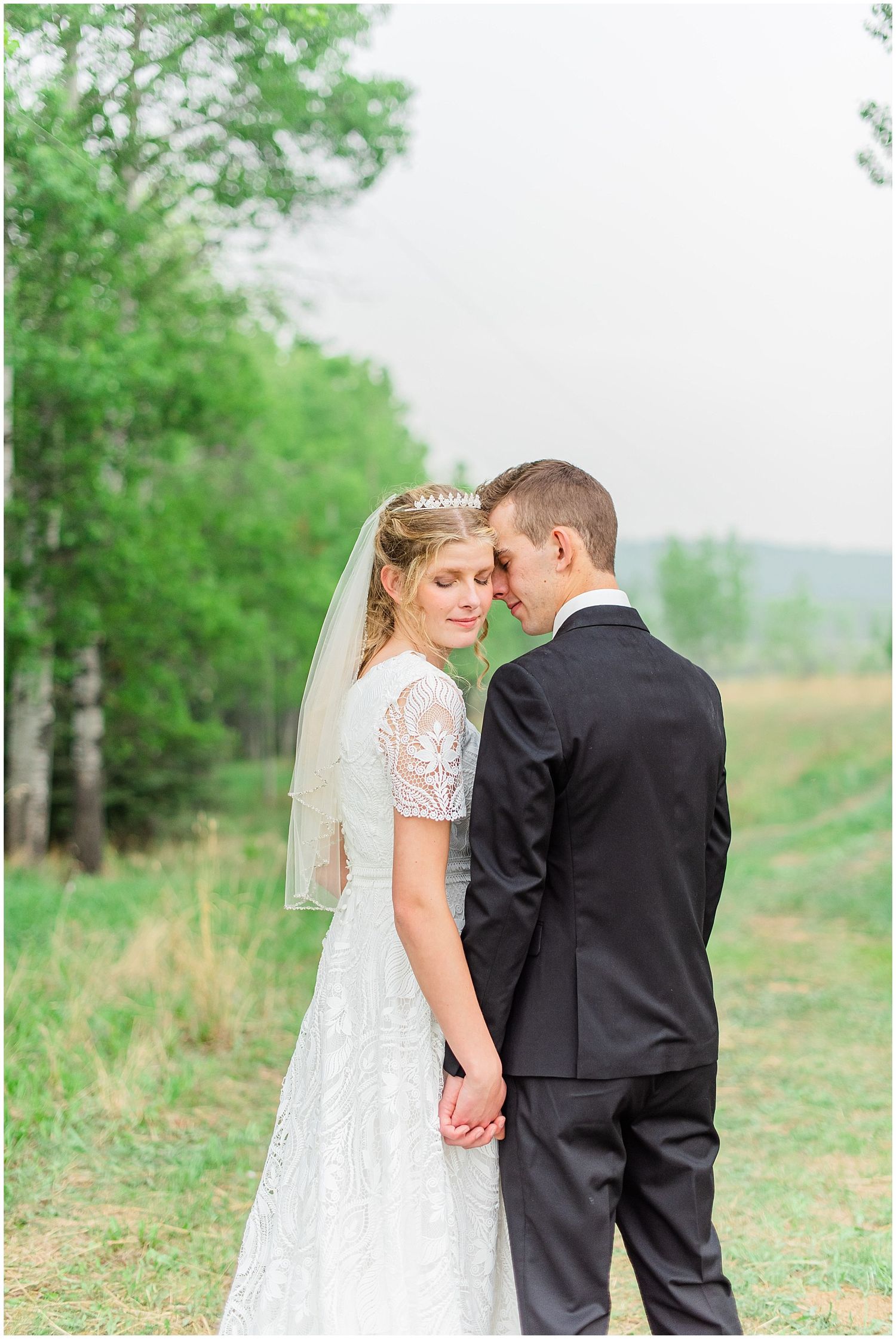 Bride and groom hold hands facing each other in a lush green field, bride wearing lace gown and veil with tiara.