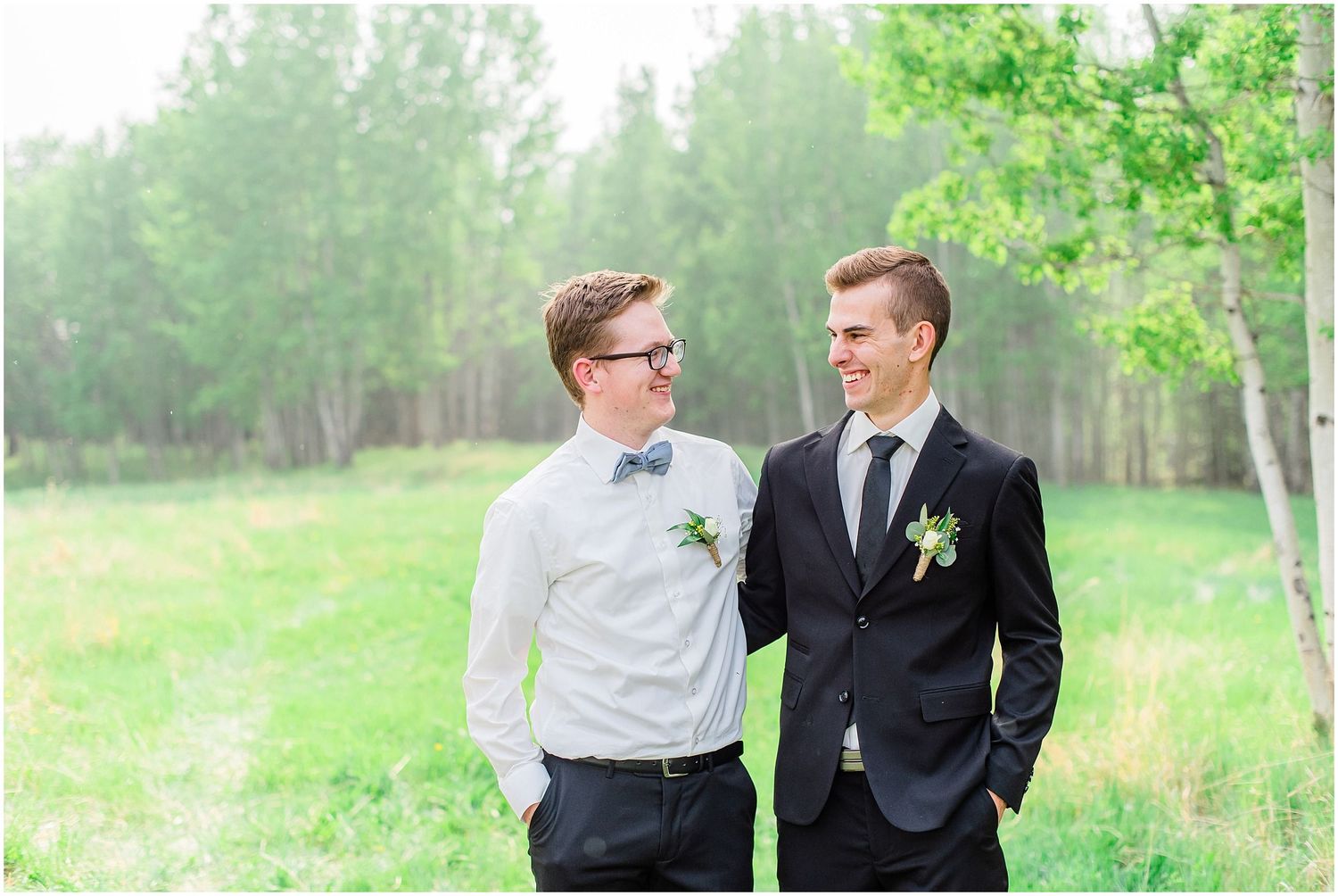 Two grooms share a smile outdoors in a lush green field, one in a black suit and one in a white shirt with bow tie.