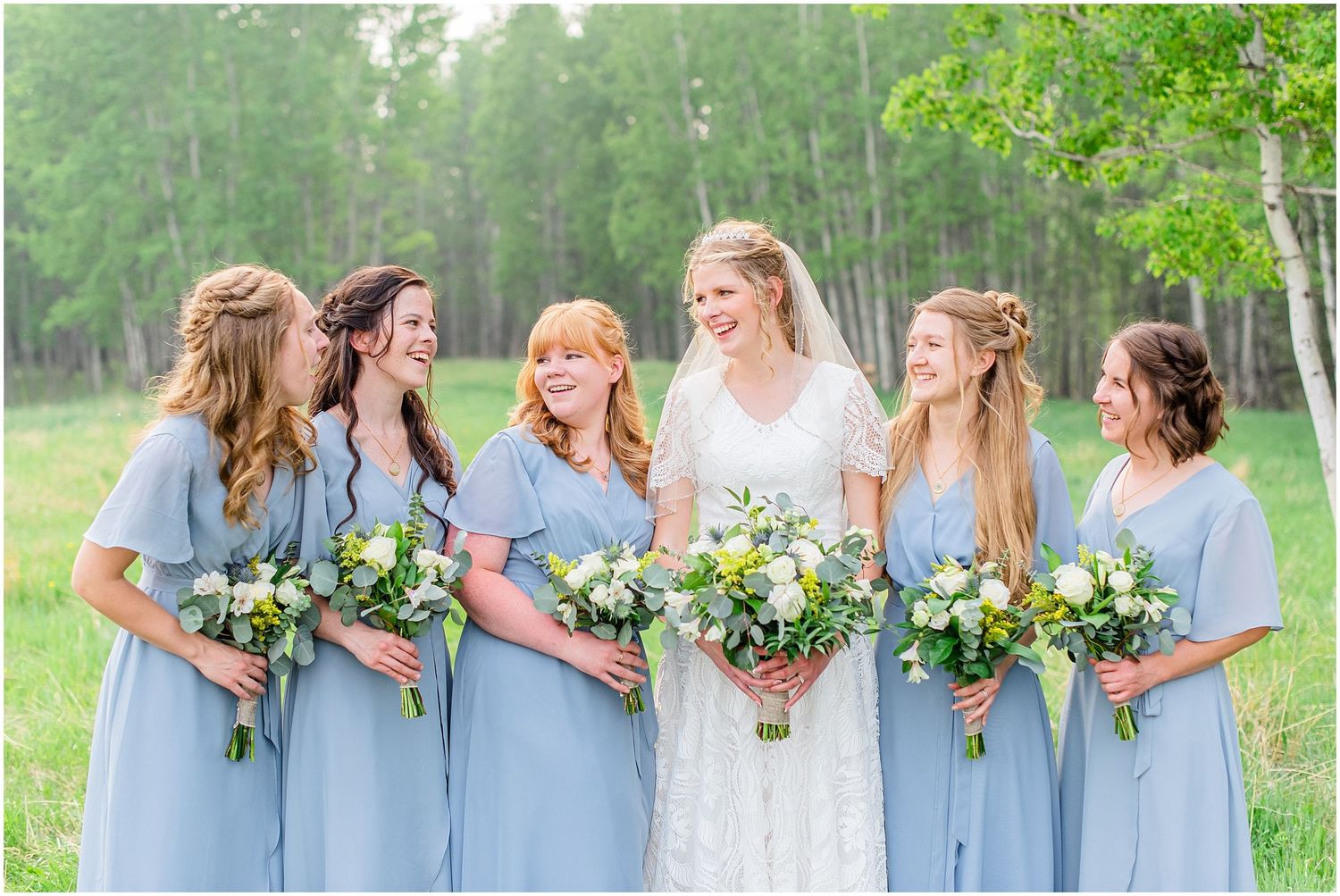 Bride in lace gown smiling with five bridesmaids in dusty blue dresses holding white floral bouquets outdoors.