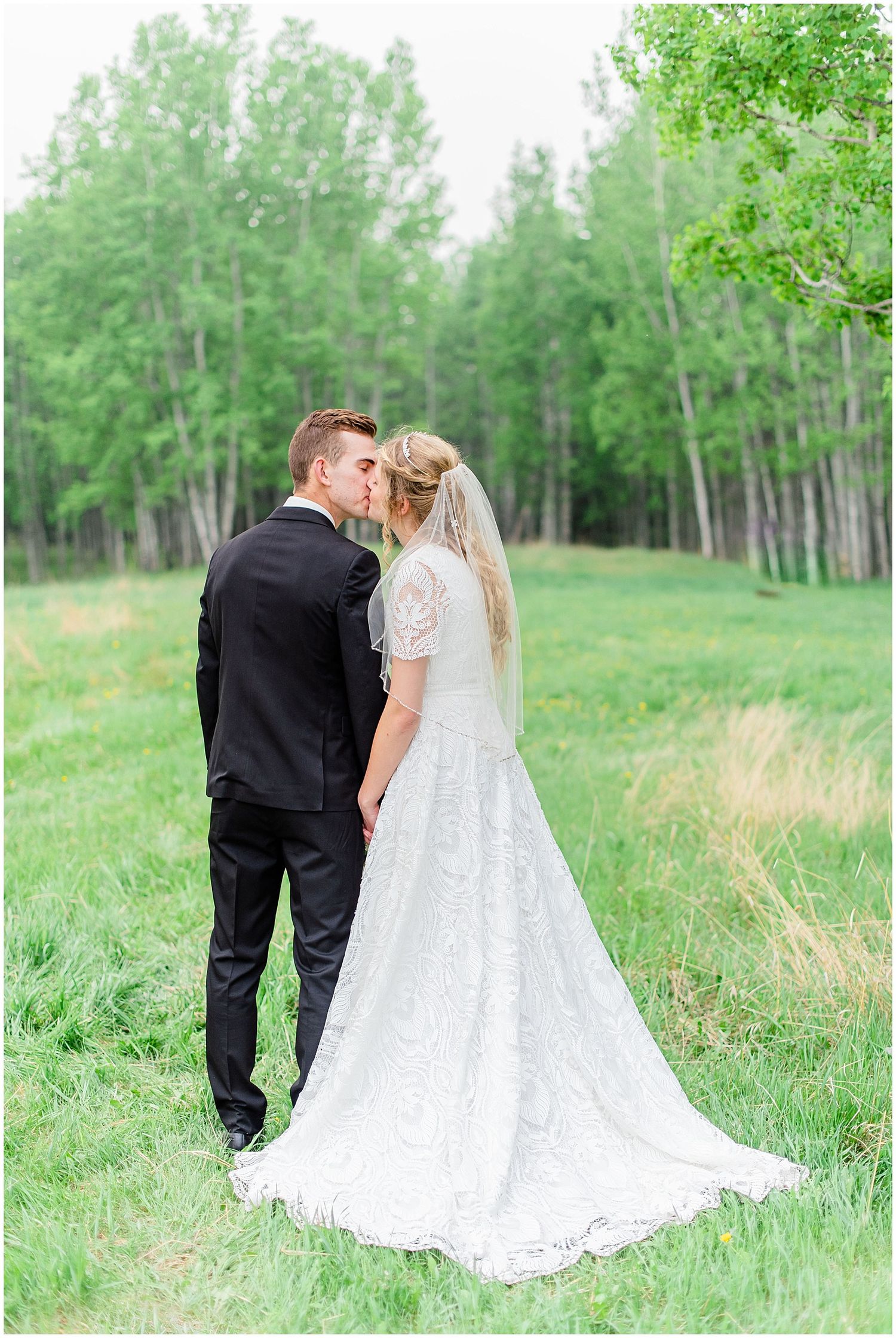 Wedding couple walks hand in hand through green meadow, bride in elegant lace gown with long train, groom in black suit.